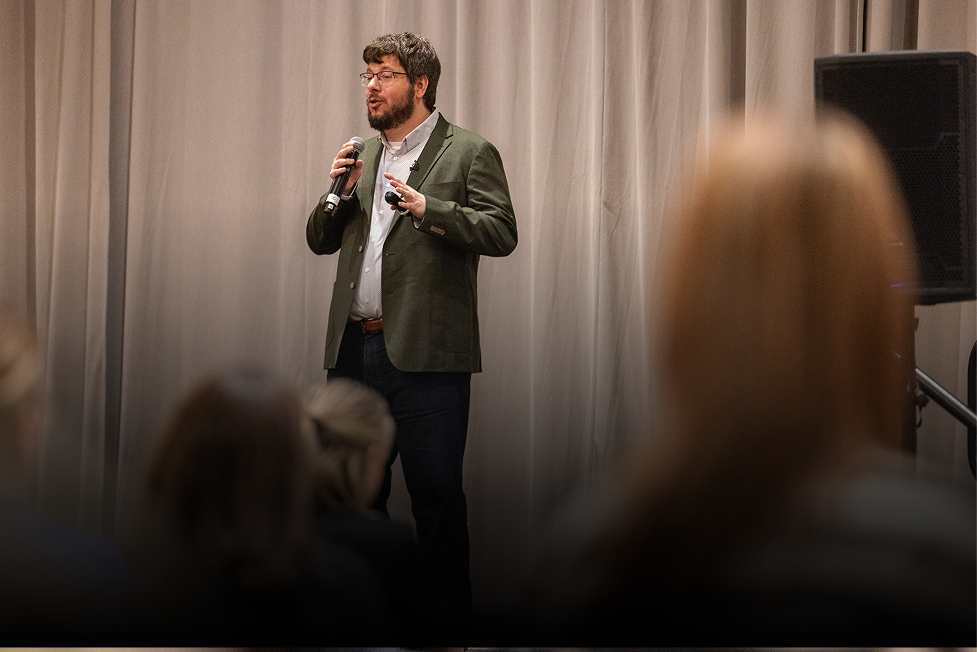 A man in a green blazer and glasses speaking into a microphone at a presentation or event.