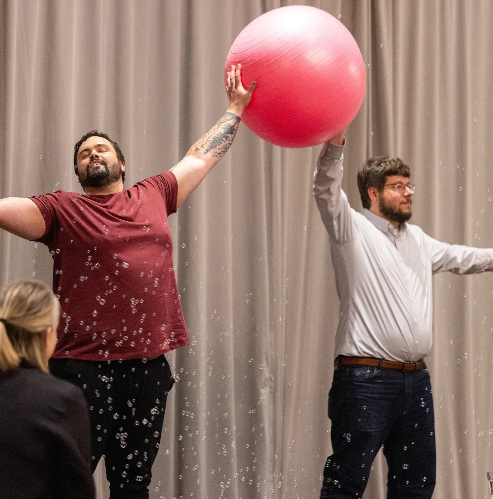 Two men are holding a large red exercise ball above their heads during an event, with a gray curtain in the background.