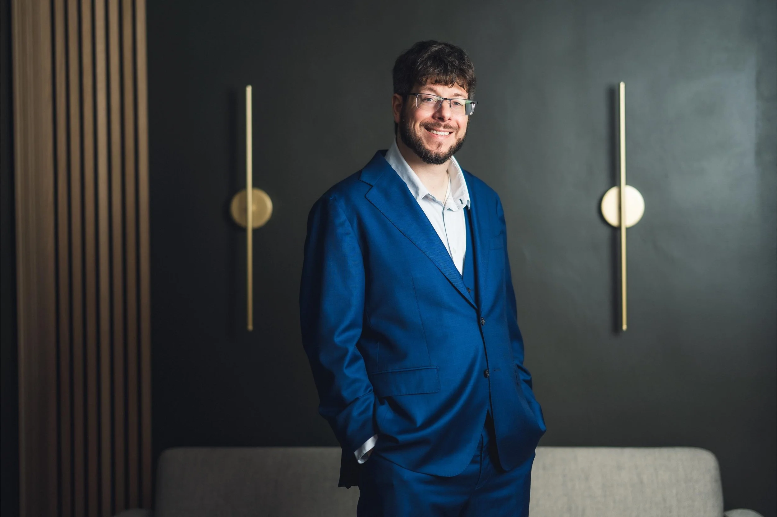 A man with brown hair, a beard, and glasses wearing a blue suit and white shirt, smiling while standing indoors against a dark wall with vertical gold and wood accents.