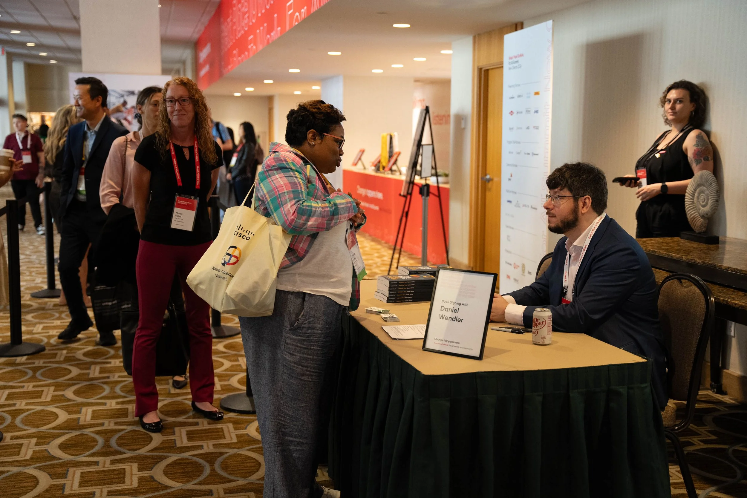People waiting in line at a book signing event at a conference. One person is sitting at a table signing a book, with a sign in front. Others stand with conference badges around their necks, engaging with the event.