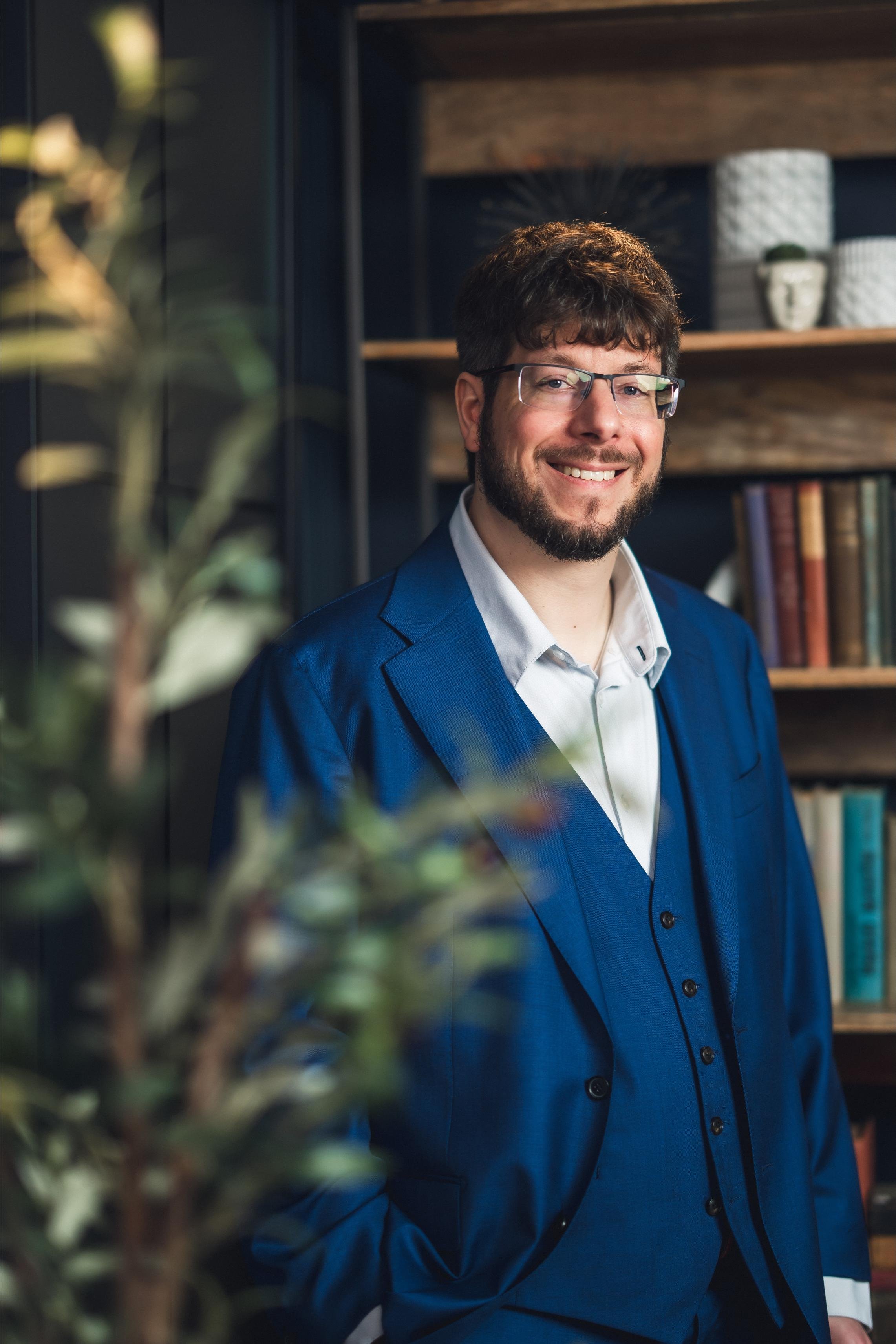 Portrait of a man with glasses, brown hair, and beard, smiling in a blue blazer, standing in a room with wood shelves and books in the background.