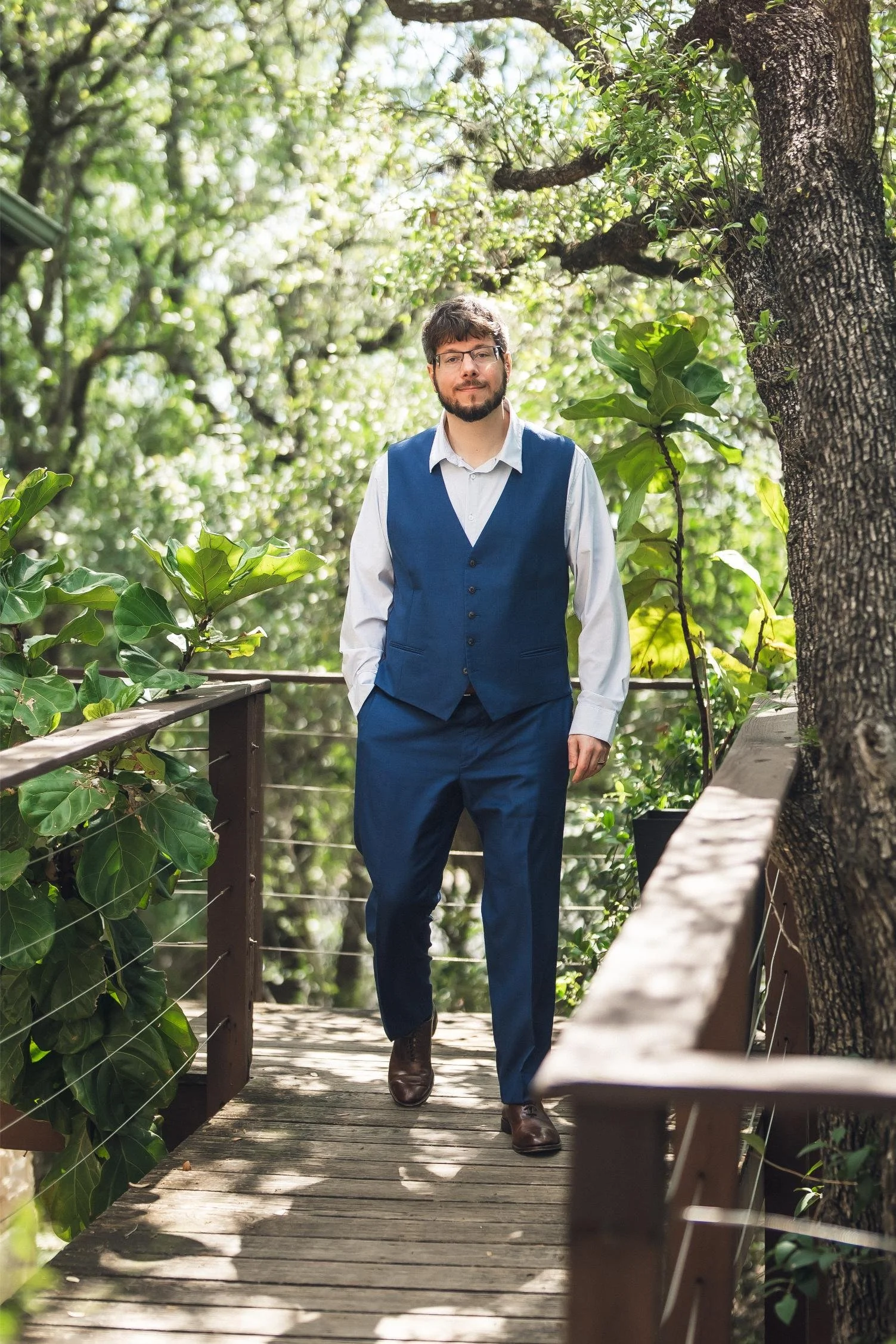 A man walking on a wooden pathway surrounded by lush green trees and plants, wearing a white shirt, blue vest, and blue pants, with glasses and a beard.
