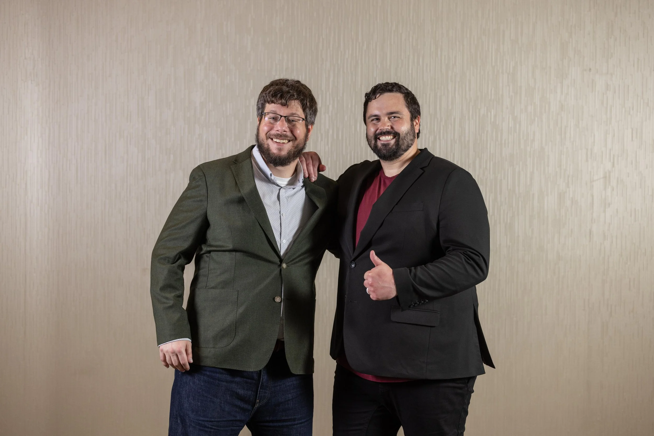 Two men in suits smiling and posing together against a plain light-colored background, one with an arm around the other's shoulder.