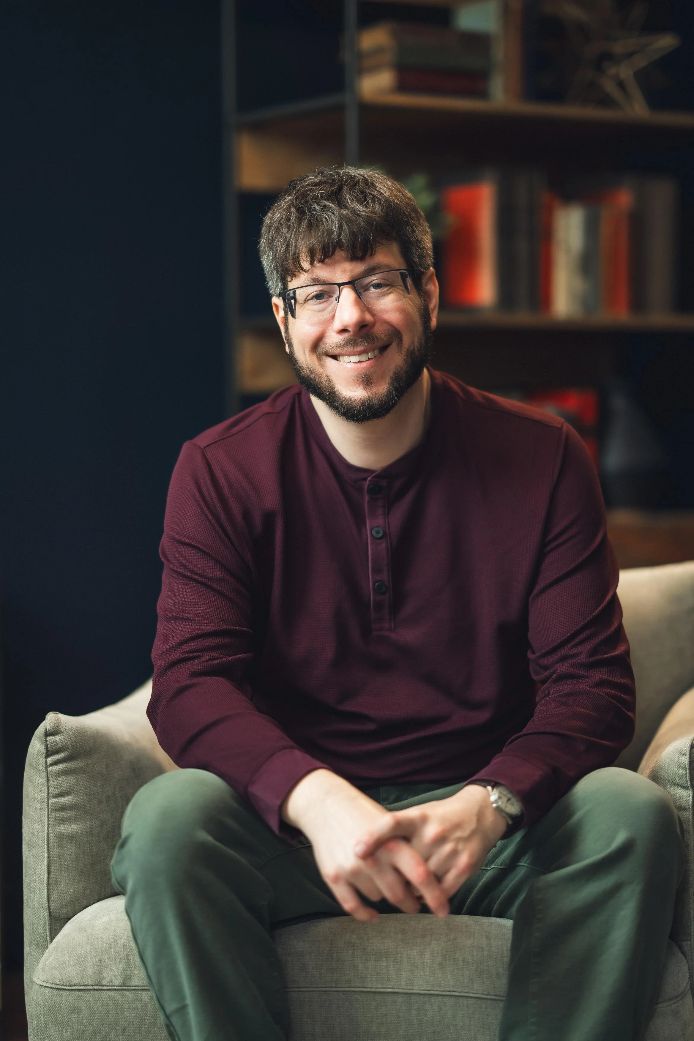 A man with glasses and a beard smiling, sitting on a light-colored armchair in a room with a dark wall and a background of bookshelves with books.