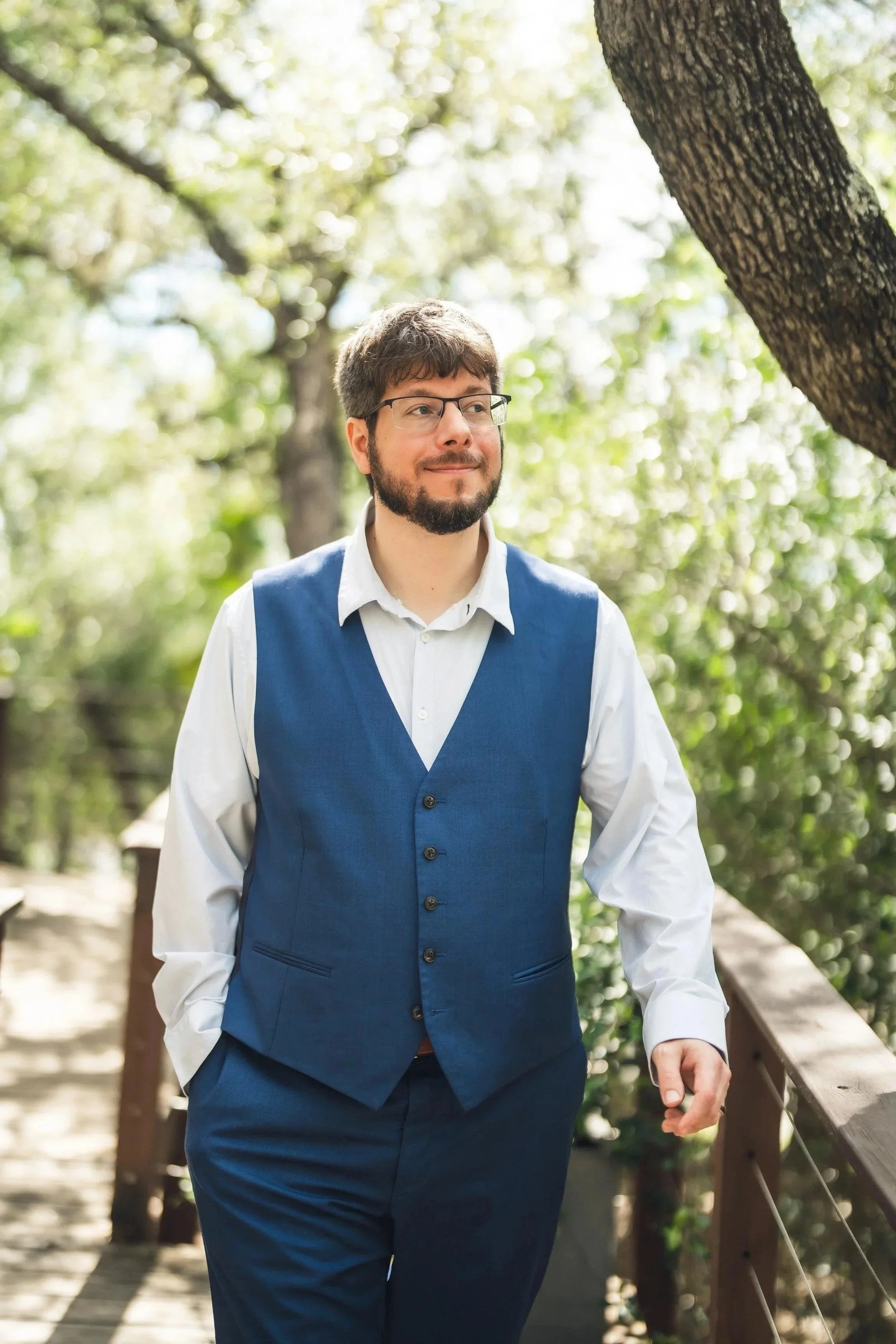A man in formal attire, wearing glasses and a blue vest over a white shirt, walking on a wooden bridge in a lush, green park.