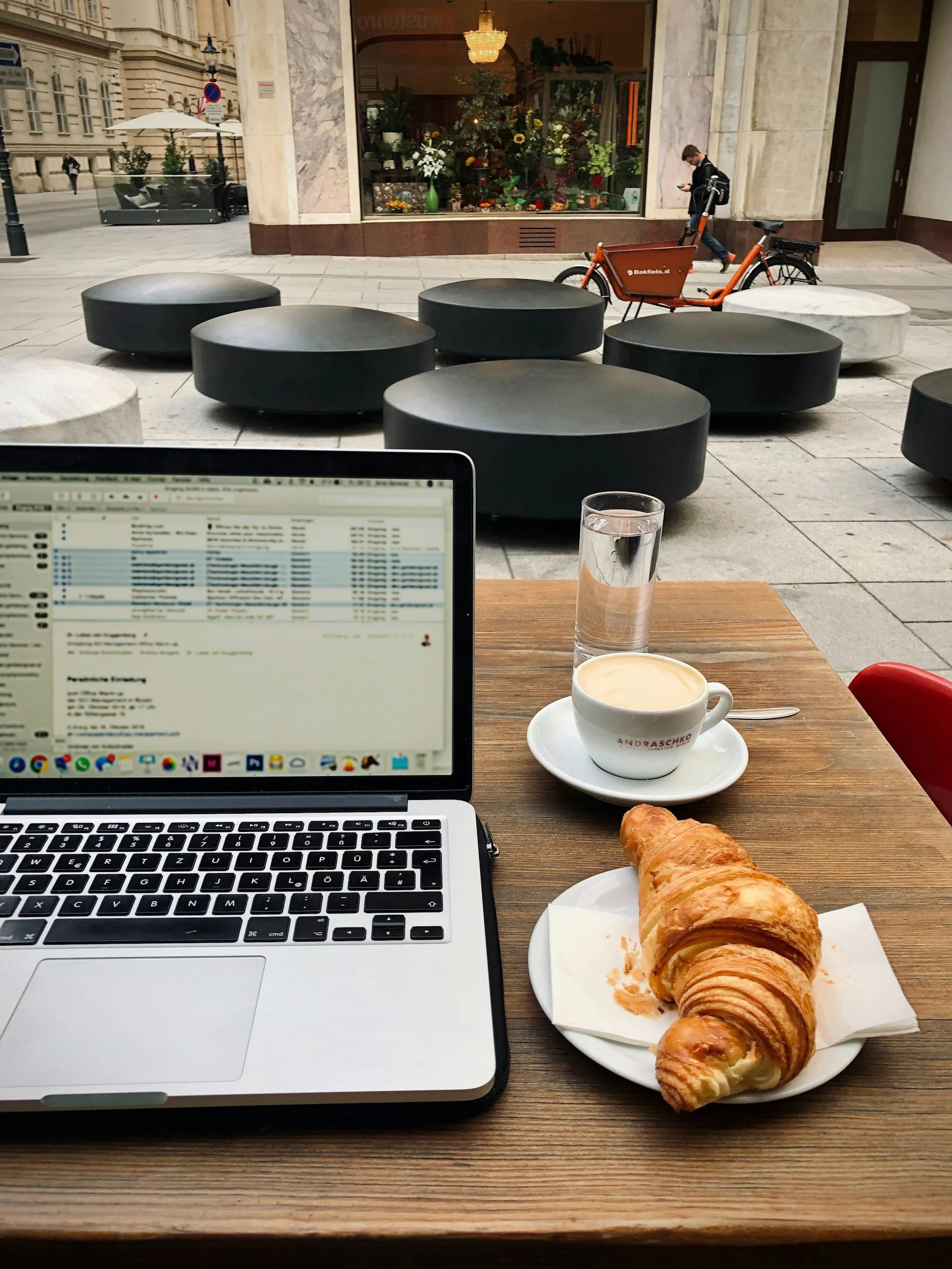 A table set outside a cafe with a laptop, a glass of water, a cup of coffee, and a croissant. In the background, there are modern urban street furniture and a storefront with flowers and plants inside, and a person walking with a backpack next to an orange bicycle.