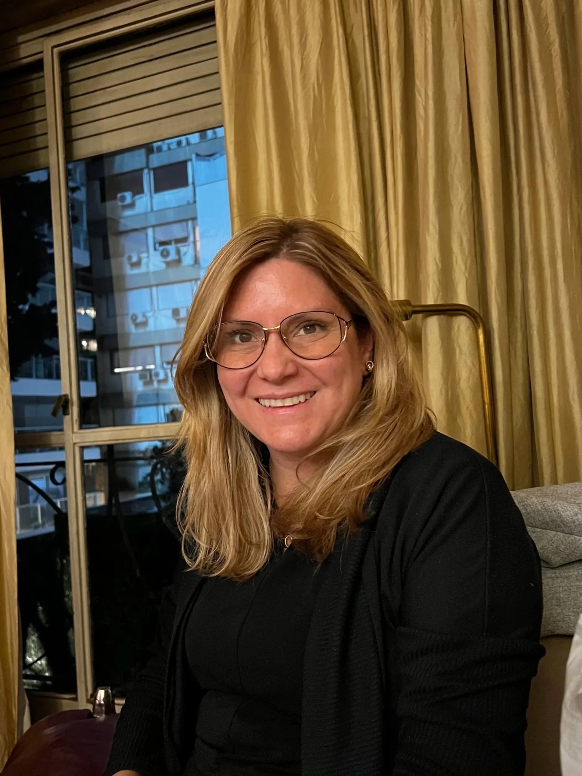 A woman with shoulder-length blonde hair, glasses, and earrings smiling at the camera indoors, with gold curtains and a window showing a building with air conditioning units outside.