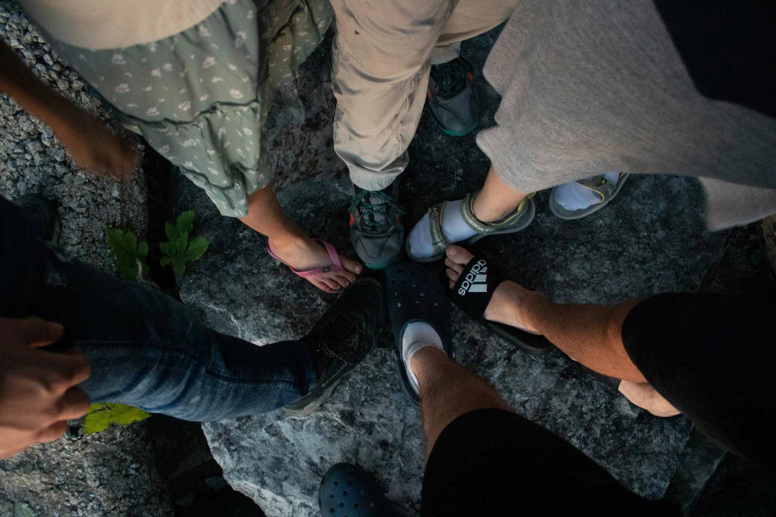 A top-down view of six people's feet standing close together on a rocky surface, showing various footwear including sneakers, sandals, and Crocs.