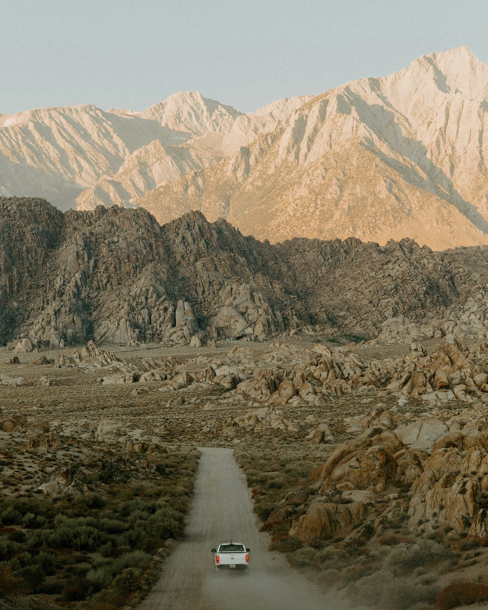 A white pickup truck driving on a dirt road through a barren desert landscape with rugged rocky hills and towering mountains in the background under a clear sky.