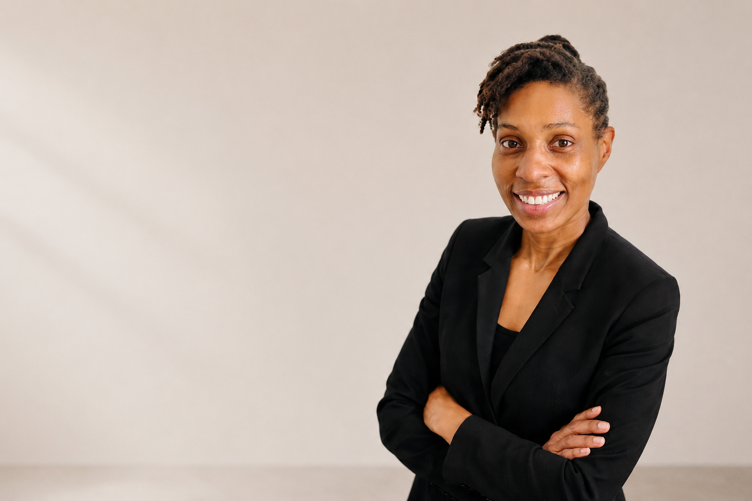 Dr. Asha Gipson, smiling with crossed arms, wearing a black blazer, standing against a plain light-colored background. Founder of Addison Leadership Advisory