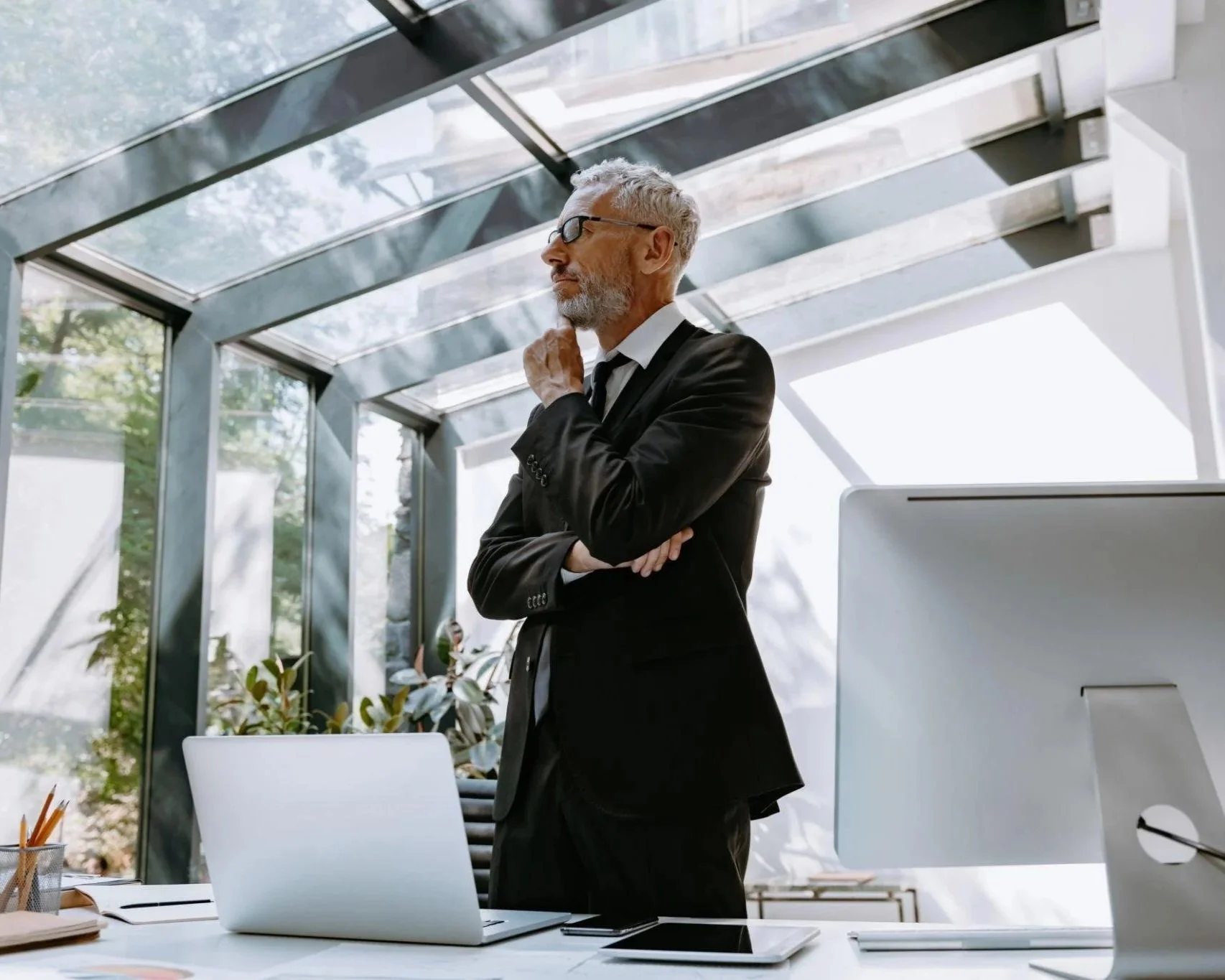 An older man in a suit and glasses standing thoughtfully in a modern office with large windows and natural sunlight.  Addison Leadership Advisory