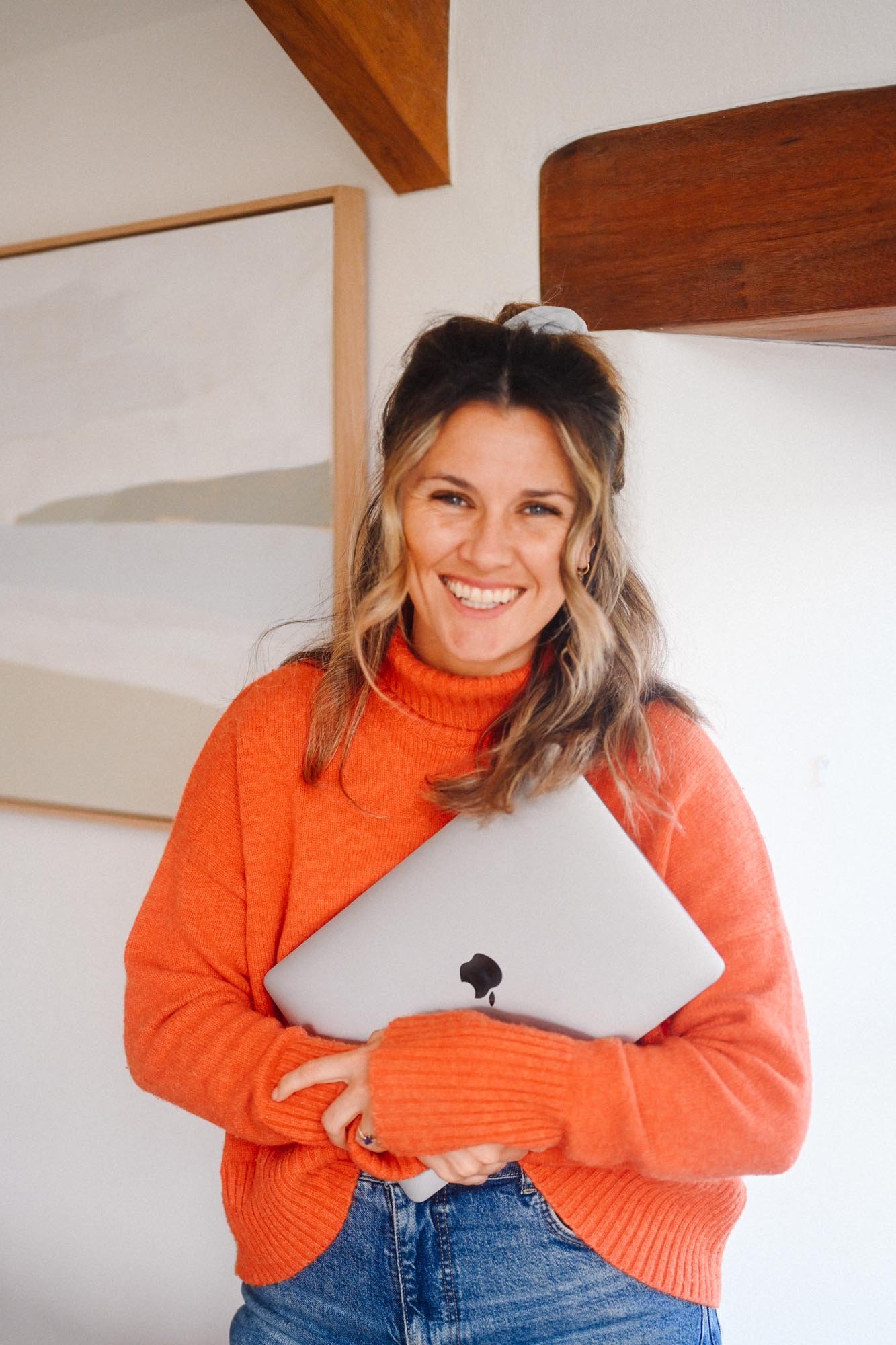 A woman with blonde hair wearing an orange sweater and blue jeans, holding a silver laptop with an Apple logo, smiling in a room with artwork on the wall