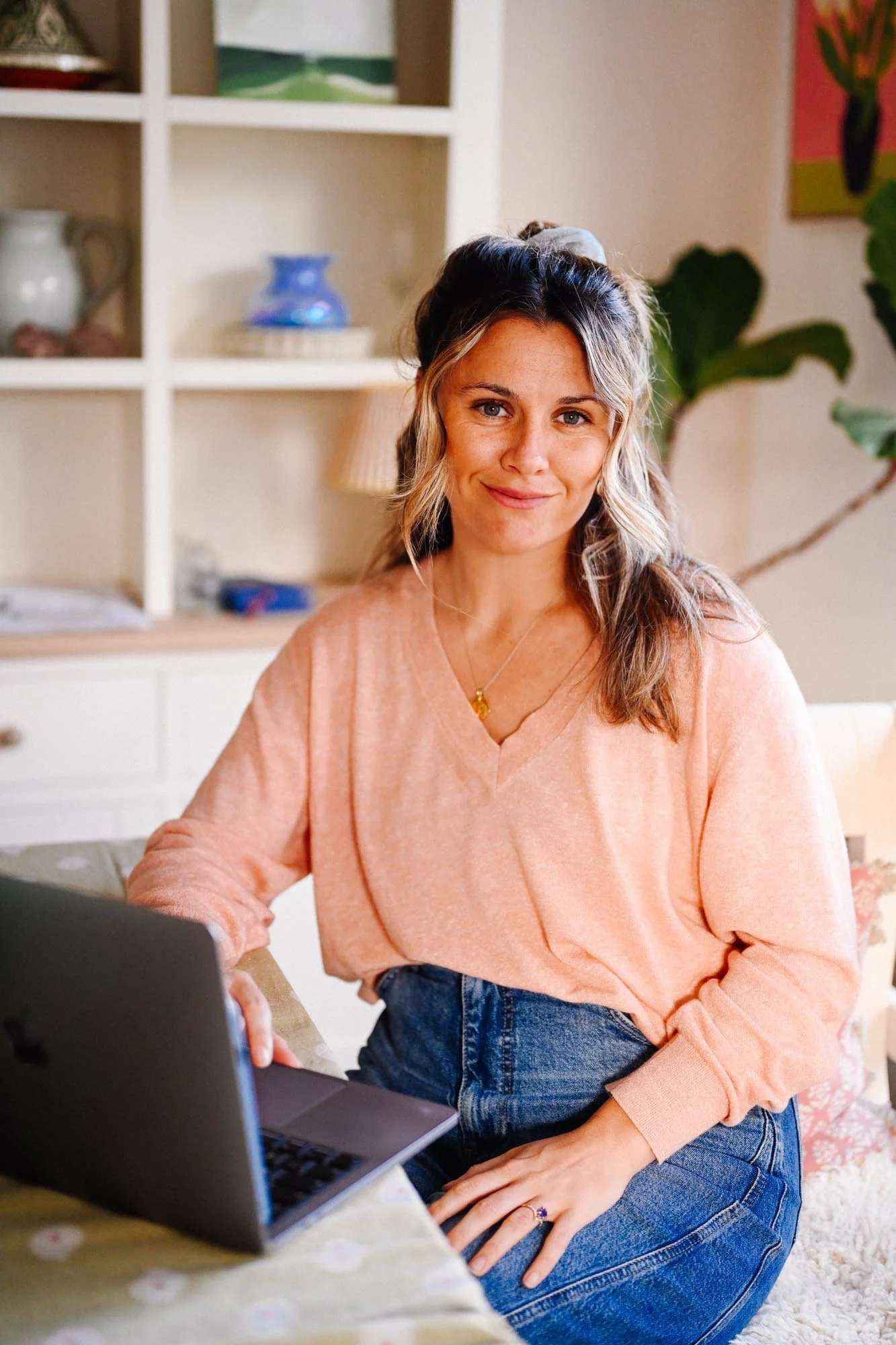 A woman sitting at a table with a laptop, smiling at the camera, in a cozy living room with bookshelf and plants in the background. Social Media Manager in Cornwall.