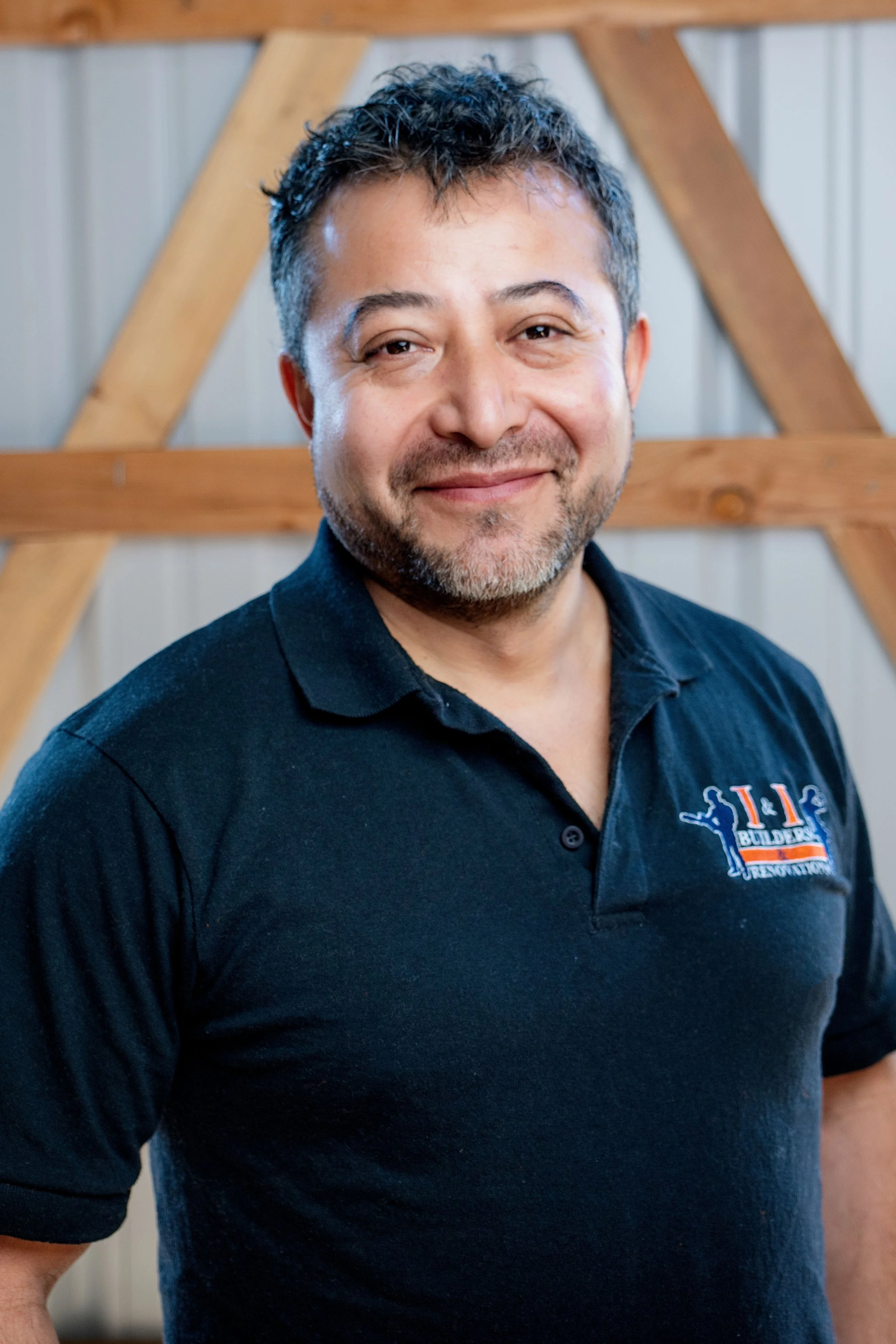 Headshot of a man with short dark and gray hair, slight beard, wearing a black polo shirt with a logo, smiling in front of a wooden background.