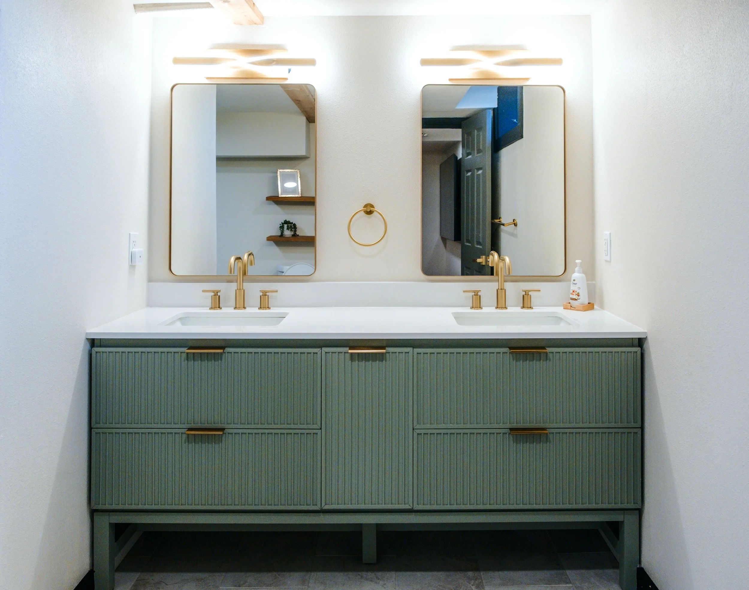 Bathroom vanity with two sinks, gold faucets, two mirrors, wooden shelves, and a hand soap dispenser.