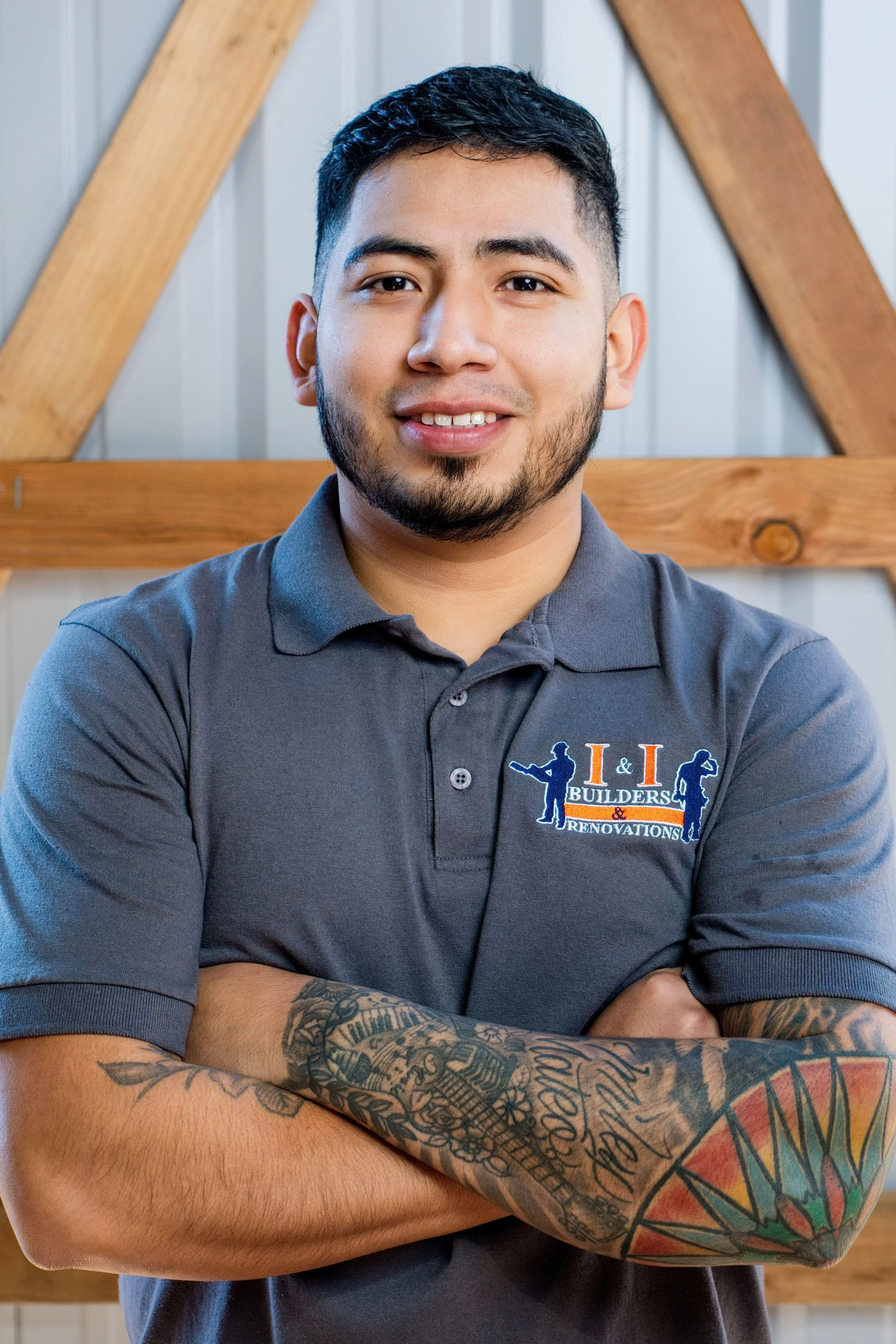 A young man with tattoos on his arms, short dark hair, and a beard, smiling with arms crossed, wearing a dark gray polo shirt with a logo on it.