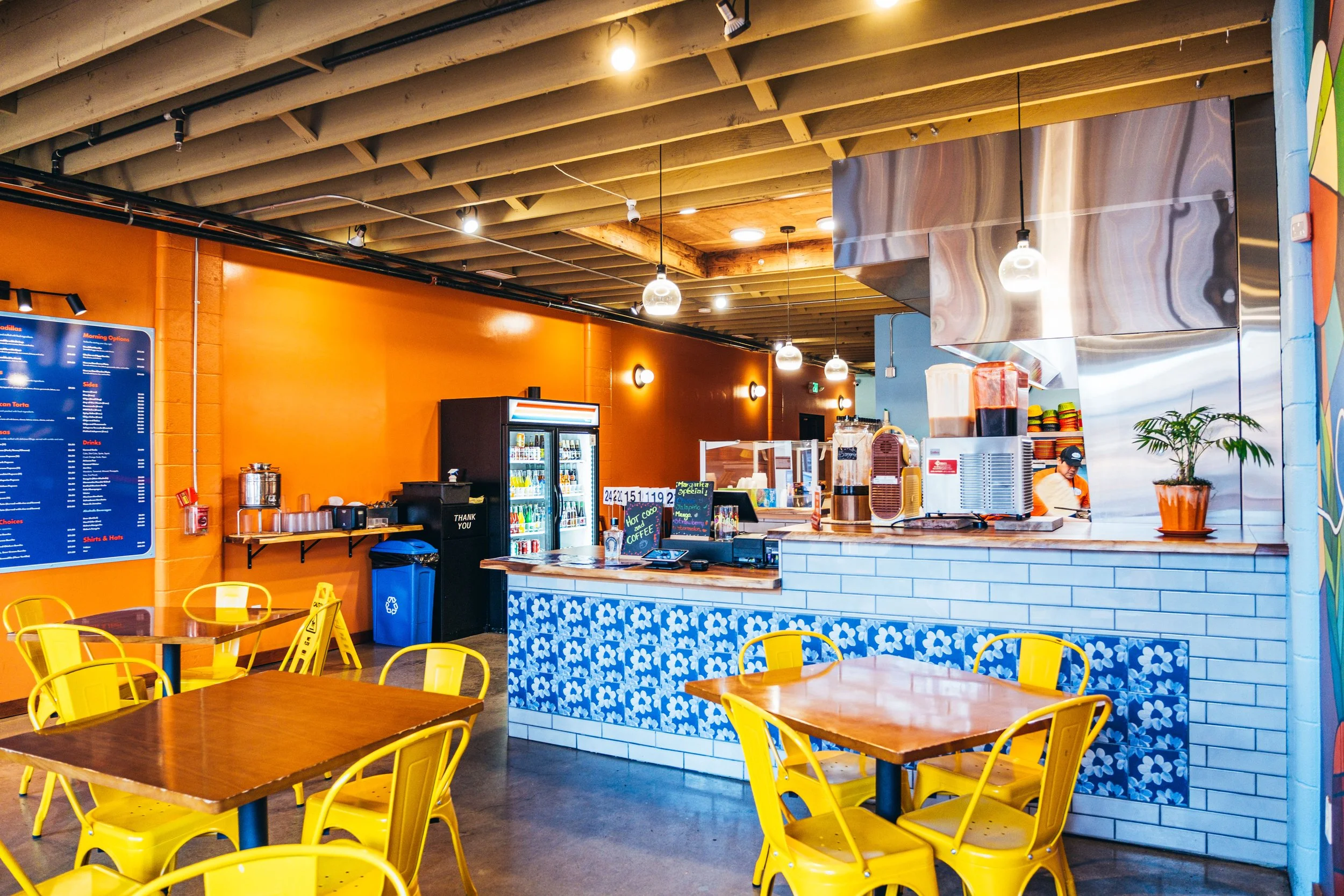 Interior of a casual eatery with yellow chairs, wooden tables, an orange wall, a menu board, a refrigerator, and a counter with a food prep area, under a wooden ceiling with exposed beams and hanging lights.