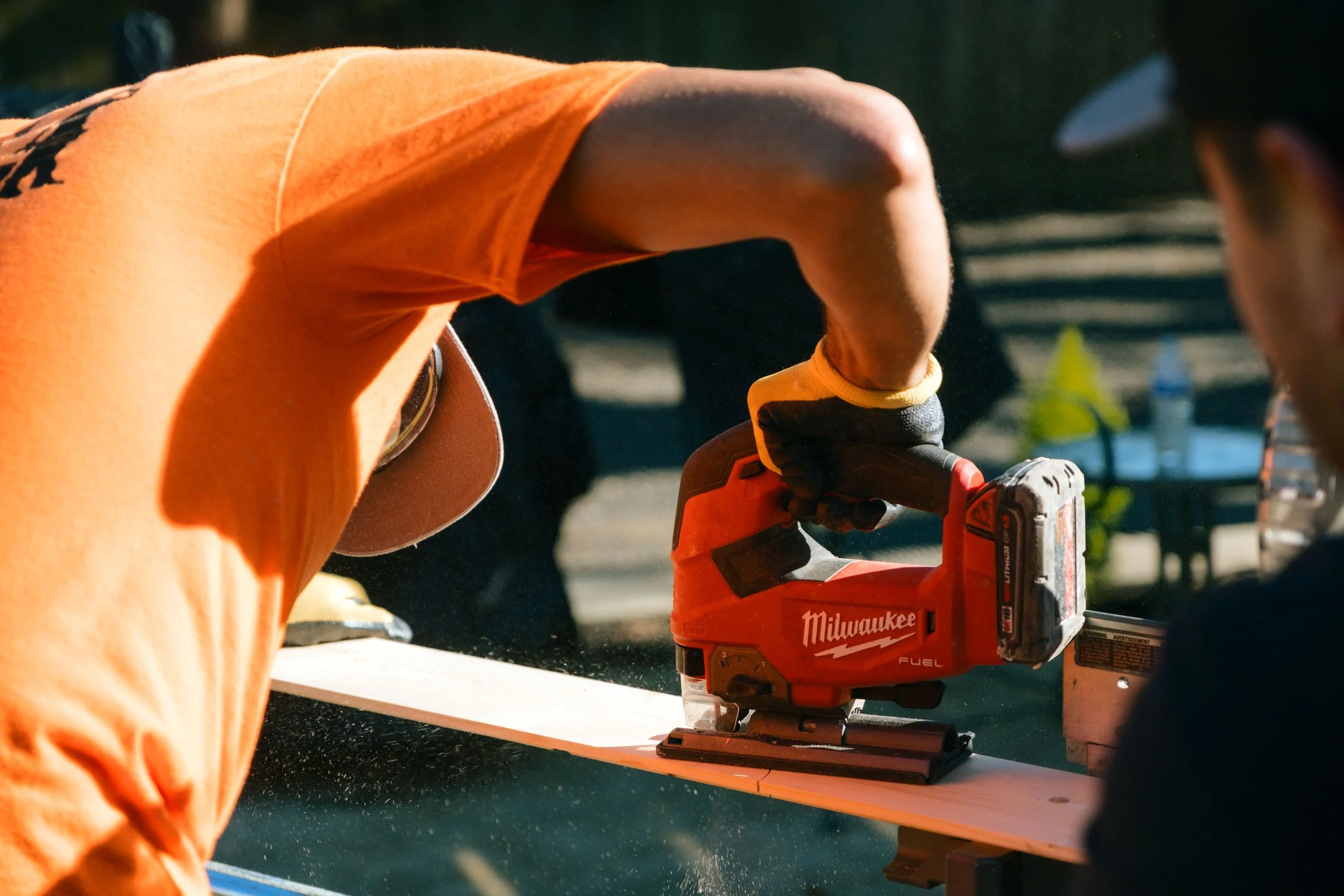 Close-up of a construction worker using an orange Milwaukee cordless power saw, wearing an orange safety vest and gloves, working outdoors.