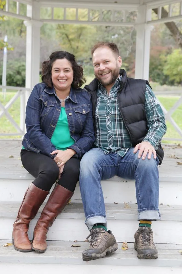 A smiling woman and man sitting on a white porch step outdoors, with green trees and a white gazebo in the background.