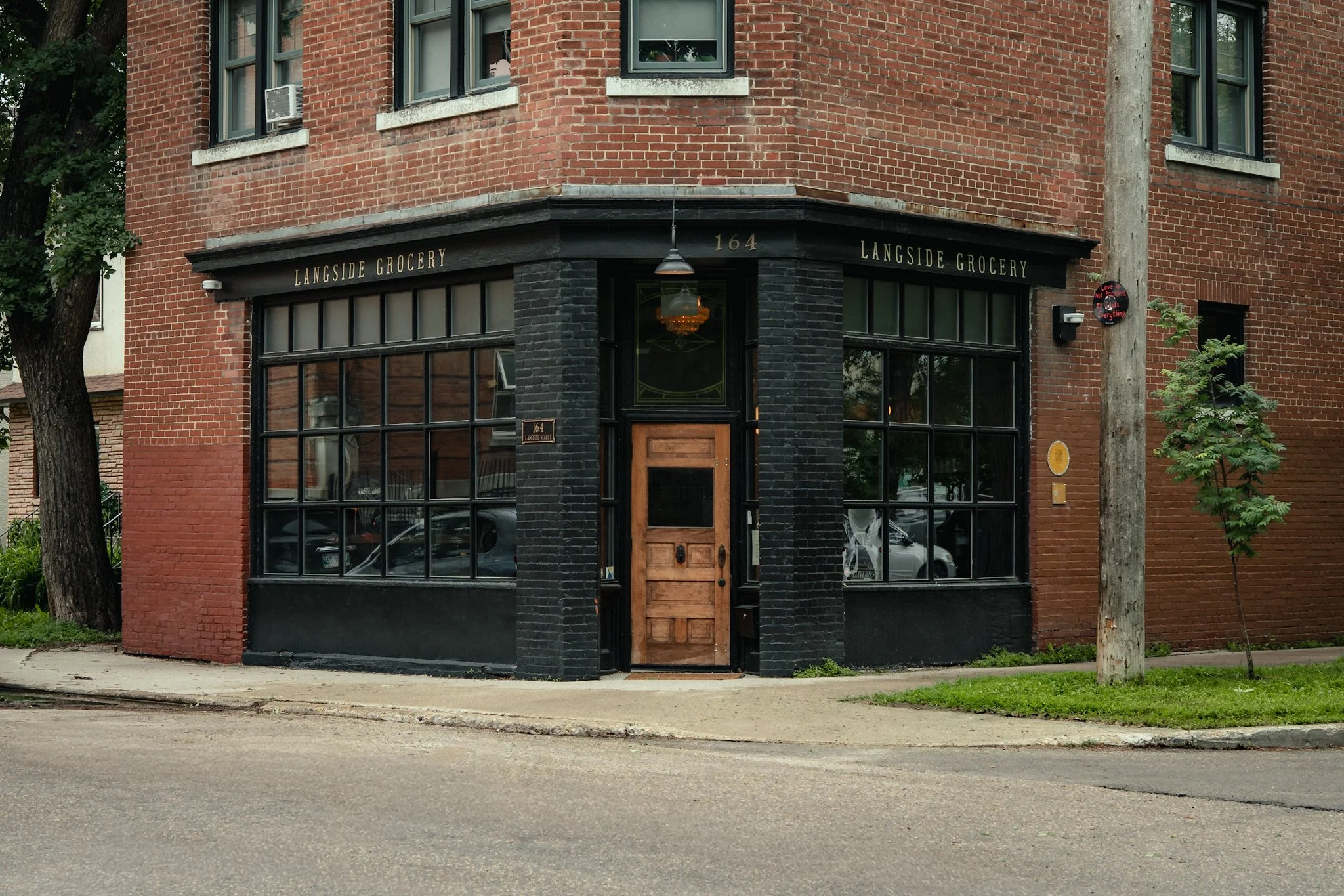A brick building with black trim on the lower floor, large windows, and a wooden door. The black trim has the text 'Langside Grocery' and the building number '164'. There are two trees in front of the building, and a sidewalk runs along the street. A small round sign with the text 'Love Not Hate Everything' is mounted on a pole near the tree on the right.
