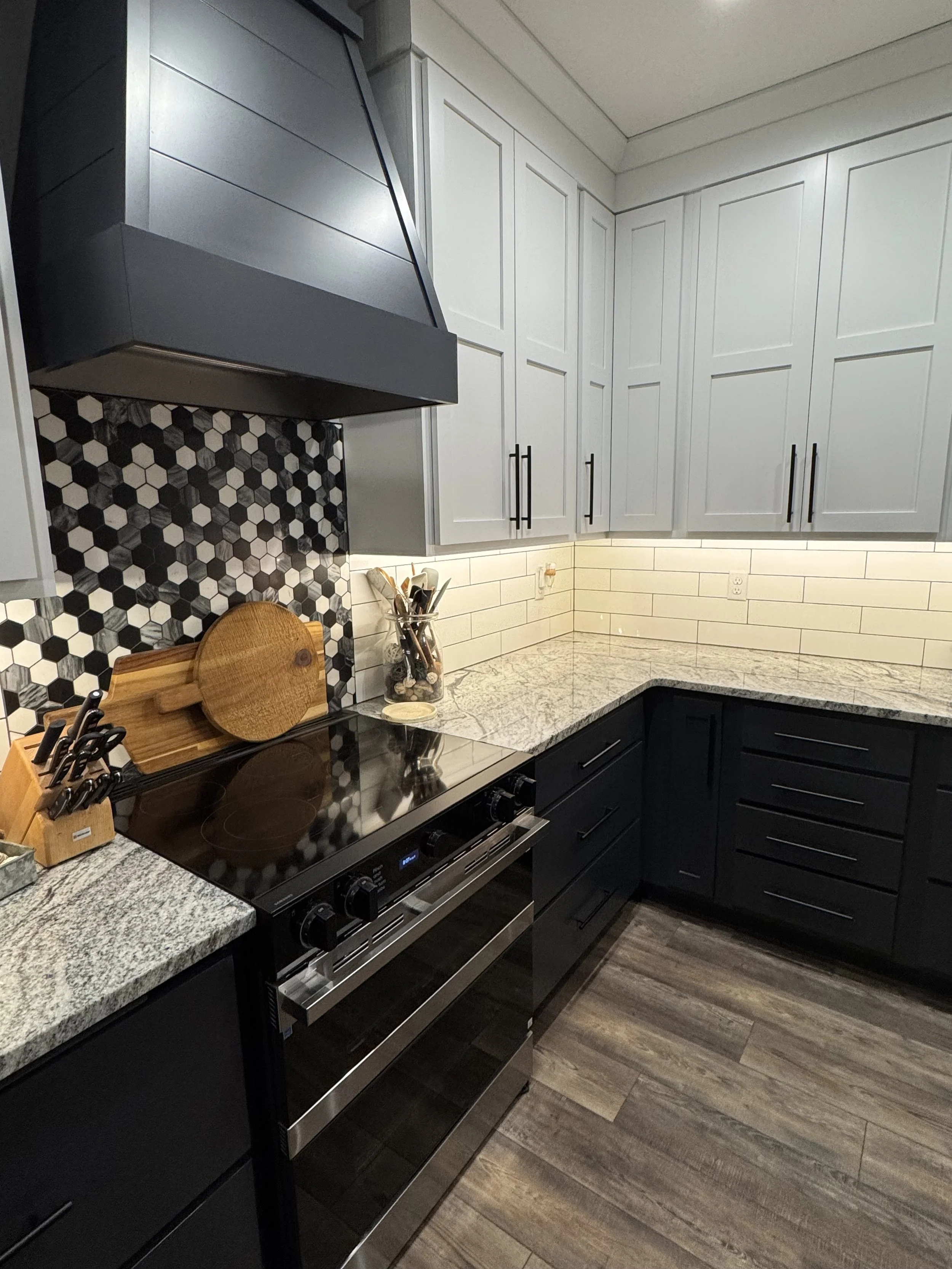 A modern kitchen with black and white cabinets, granite countertops, and a black stove. There is a hexagonal black and white tile backsplash above the stove, with a wooden cutting board, a glass jar with utensils, and a knife block on the counter.