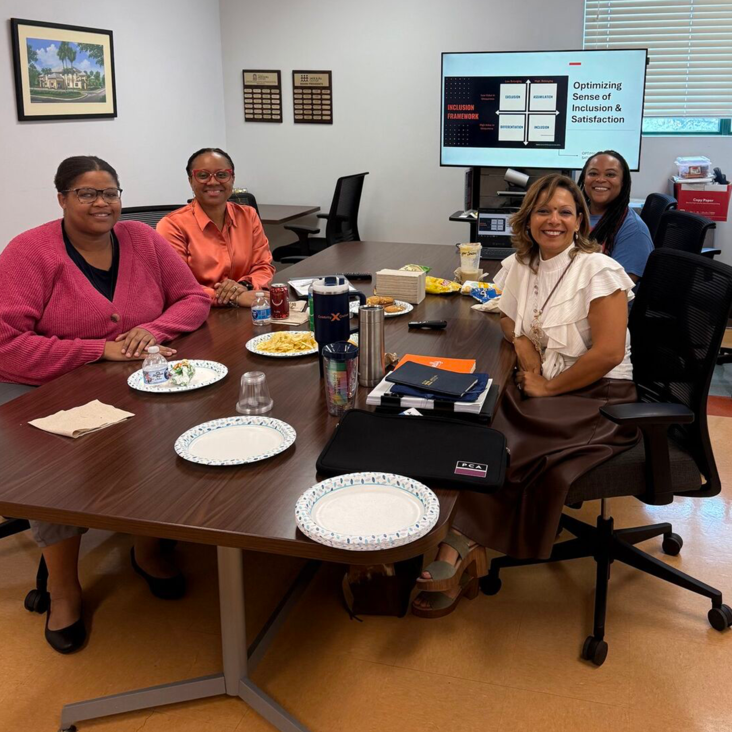 A group of five women sitting around a conference table in a room with a large screen and conference room decorations, smiling at the camera.
