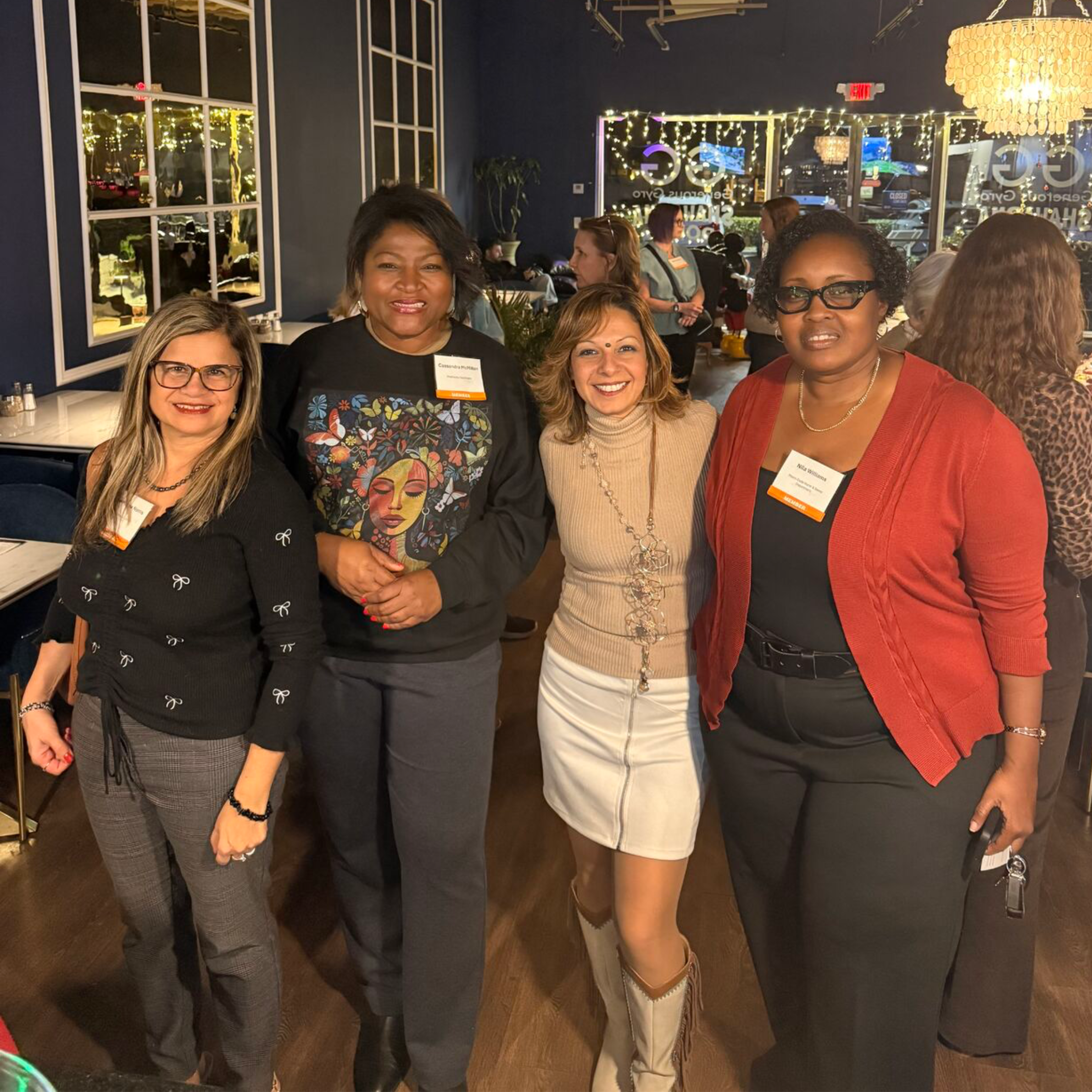 Group of five diverse women posing together indoors at a social event, with decorative lighting and large windows in the background.