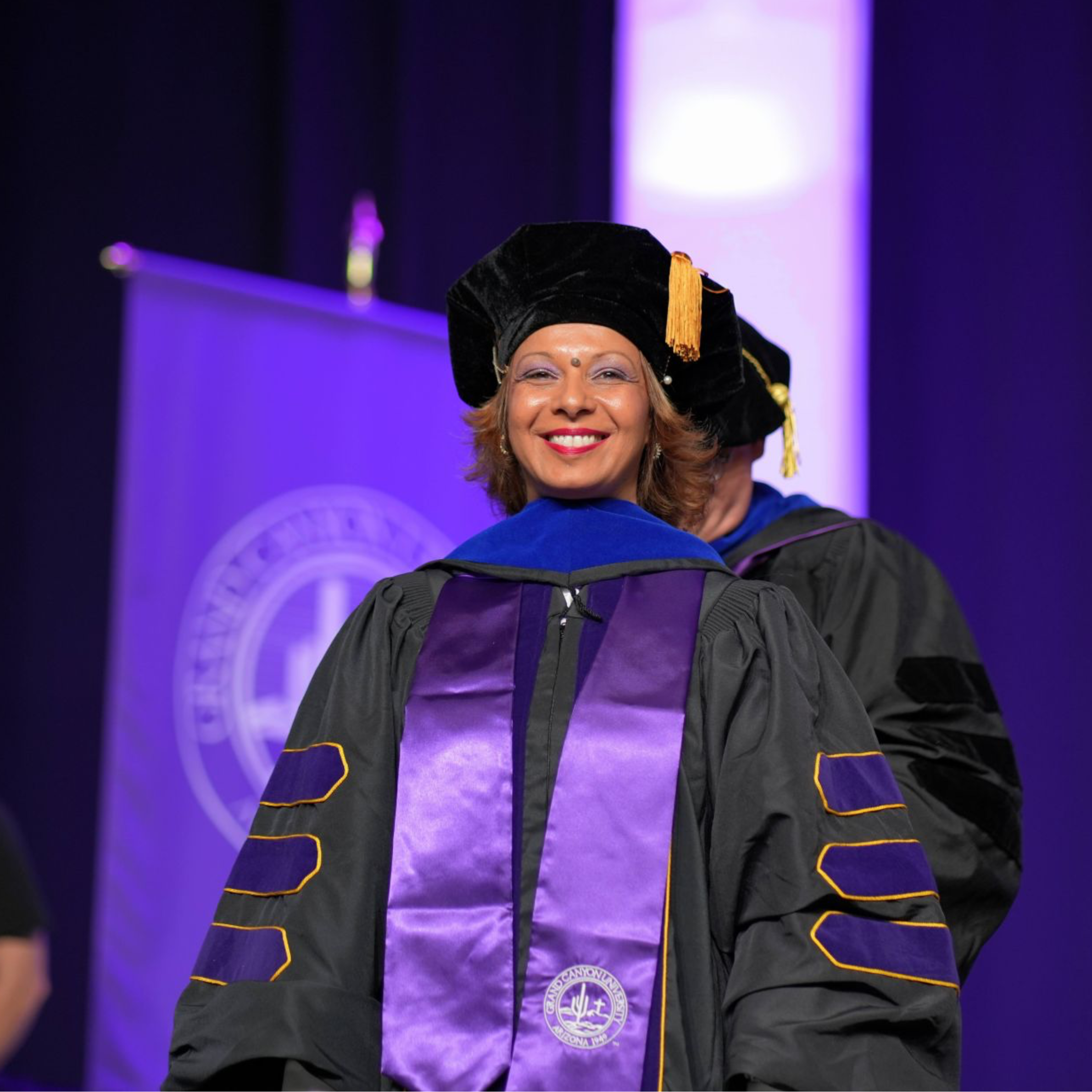 A woman wearing academic regalia, including a black cap with gold tassel, smiling at a graduation ceremony with purple and blue lighting in the background.