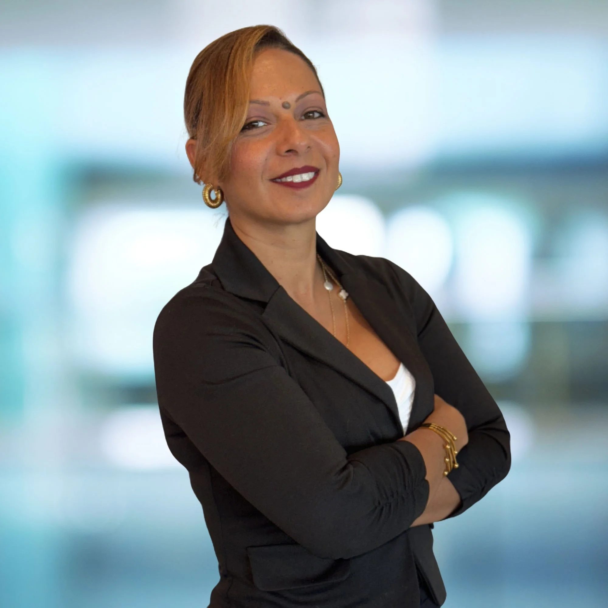 A woman with short reddish-brown hair smiling, wearing earrings and a black blazer, standing with arms crossed against a blurred light blue background.