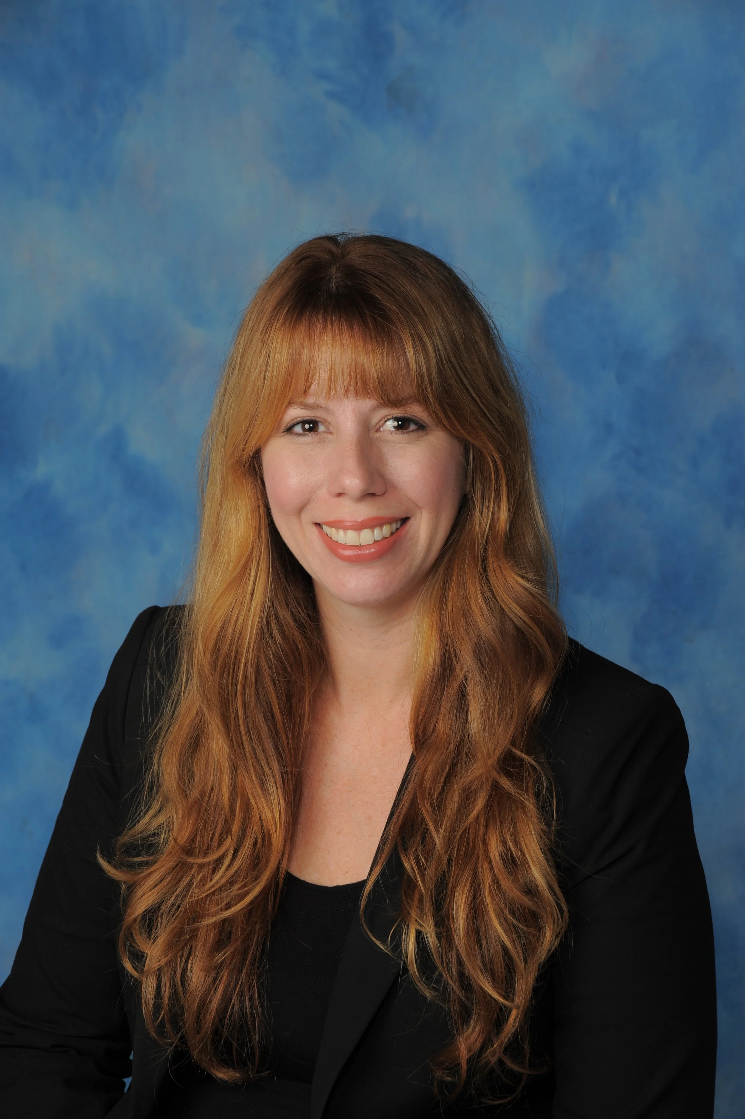 Portrait of a woman with long red hair, smiling, wearing a black blazer, against a blue cloudy background.