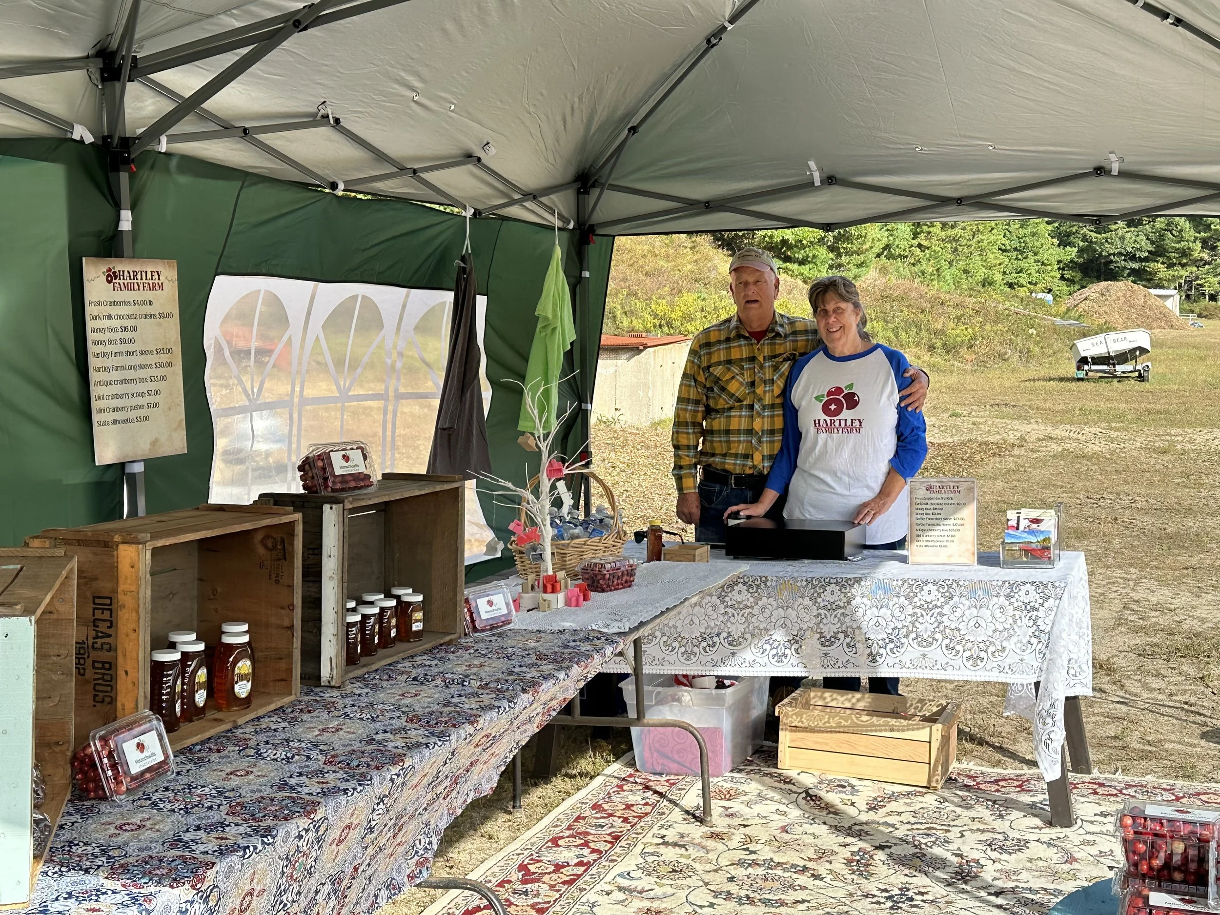 Sharon and Woody Hartley standing behind a table at the Hartley Family Farm gift shop, with jars of honey and cranberry products on the table, under a green canopy, with a sign listing various farm products.