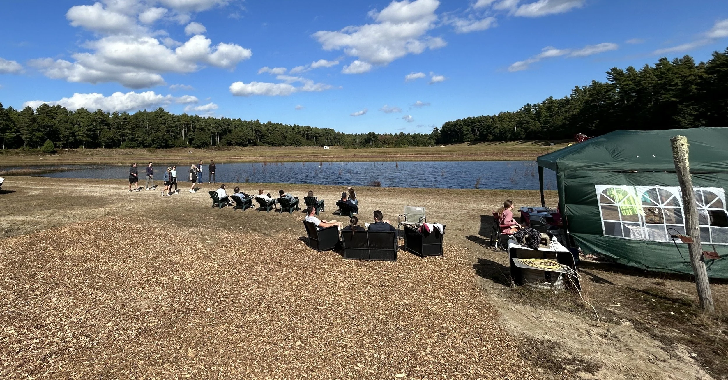 People gathered outdoors on the edge of a bog, some seated in chairs and others walking. There is a green canopy tent and chairs that is the gift shop, with a scenic background of trees and a blue sky with scattered clouds.