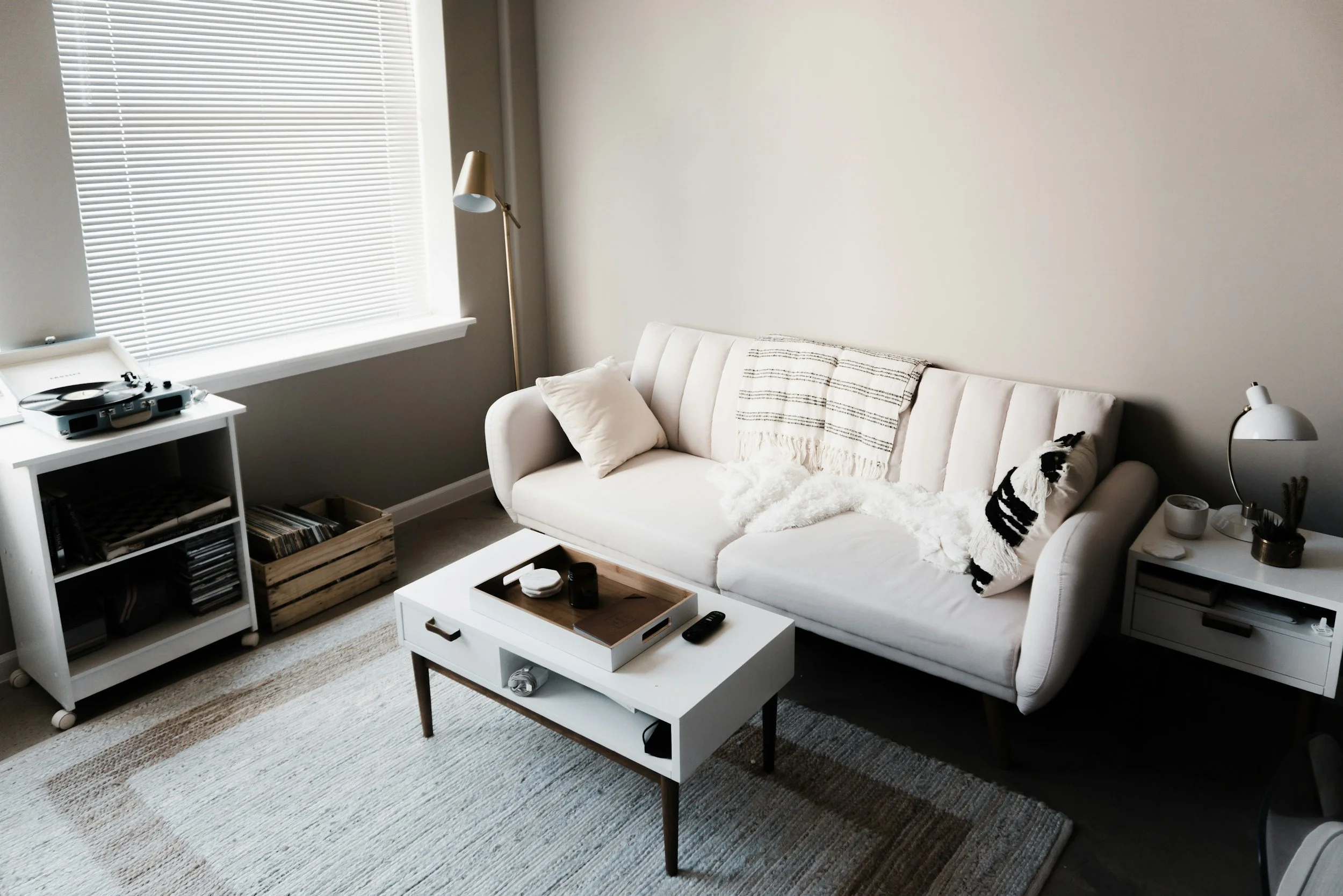 A calm, friendly room with a white couch, a matching rug, a coffee table, a window with blinds, and an accent chair.