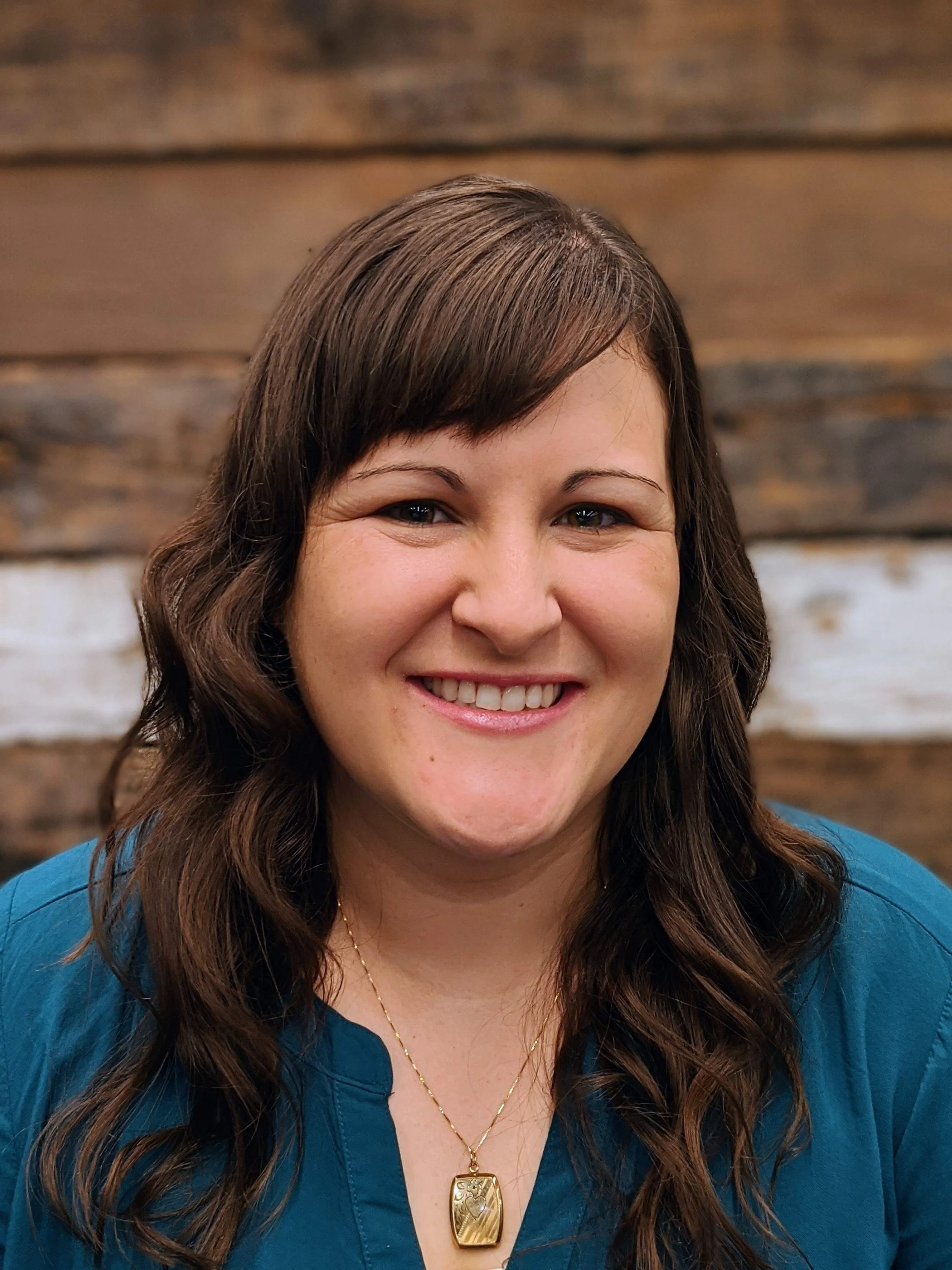 A smiling woman with long wavy brown hair wearing a teal top and a gold pendant necklace, standing in front of a wooden background.