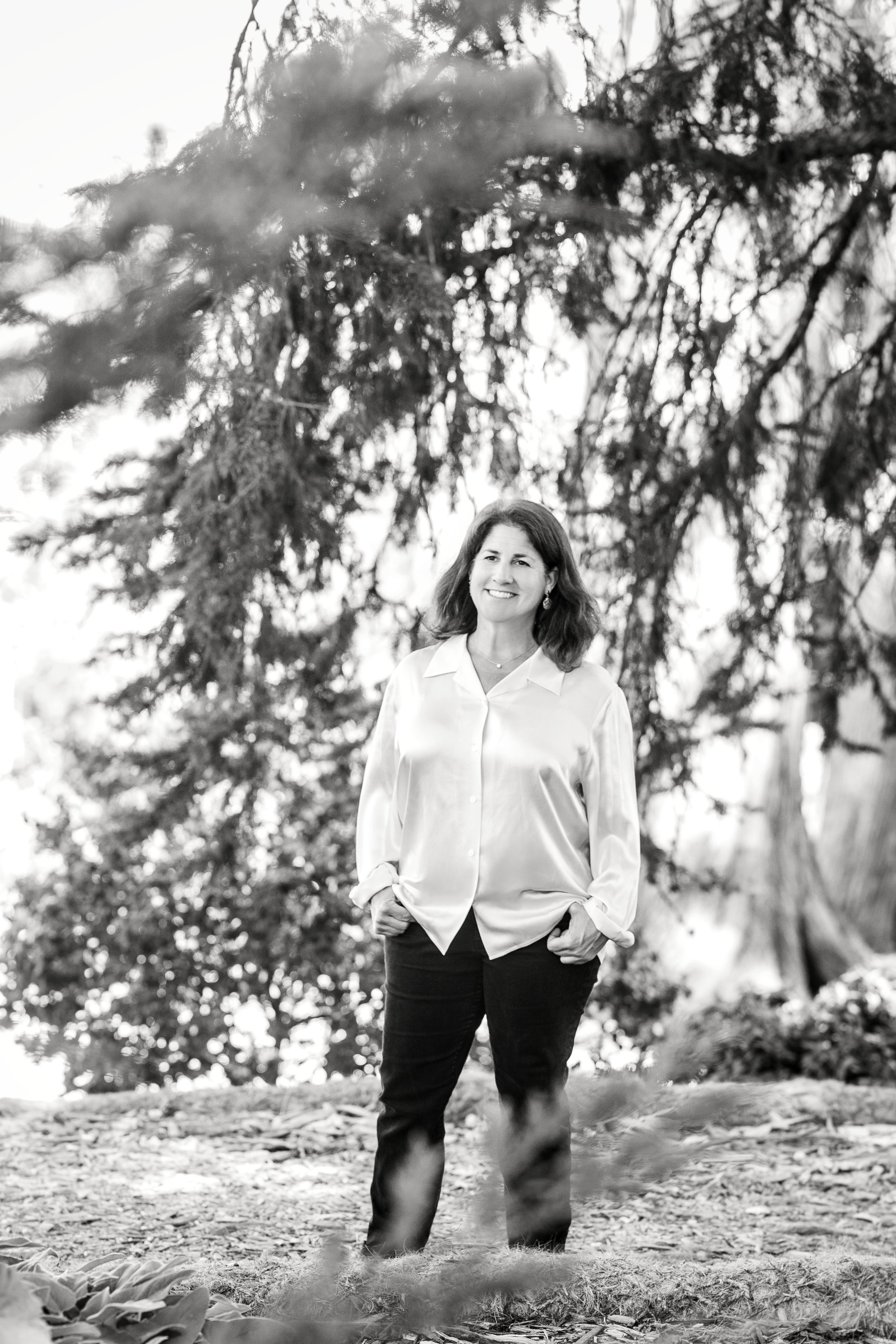 A woman standing outdoors in front of trees, smiling, wearing a white shirt and dark pants, with her hands in her pockets.