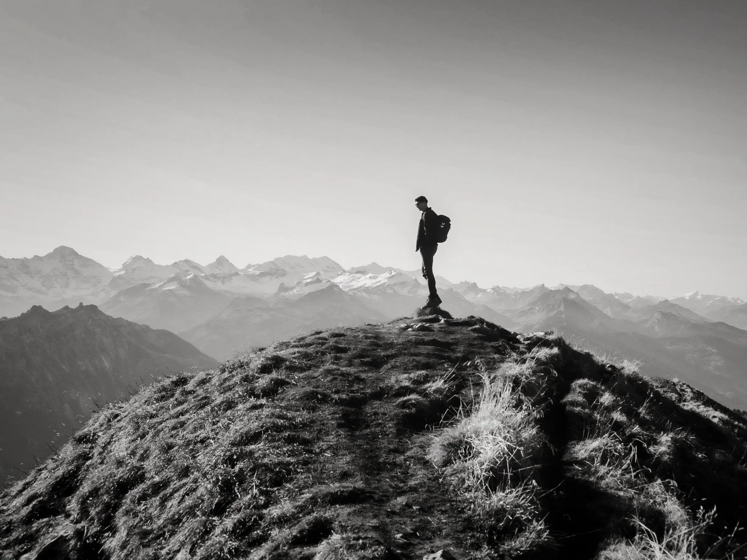 A person wearing dark clothing and sunglasses standing alone on a small hilltop, with snow-capped mountains in the background, in black and white.