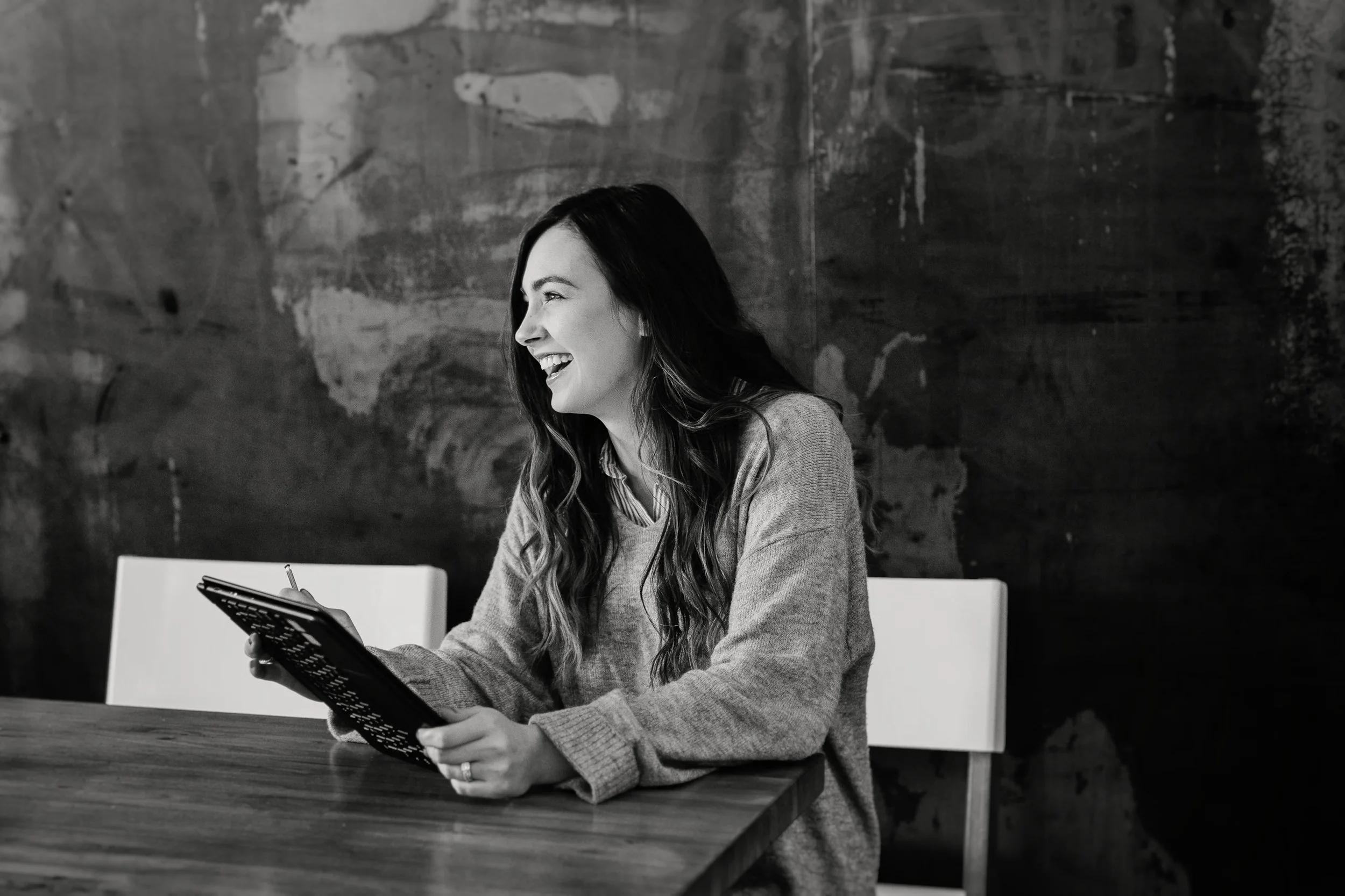 A woman with long dark hair and a striped shirt laughing while reading a tablet at a wooden table in a room with a textured dark wall.