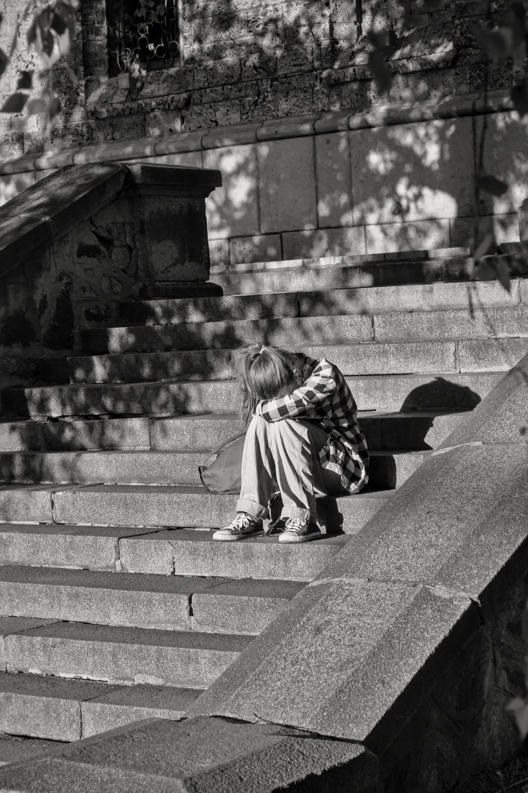 A girl with long hair sits on steps with her head resting on her knees, wearing a plaid shirt, jeans, and sneakers, with shadows of leaves cast on the steps in black and white.