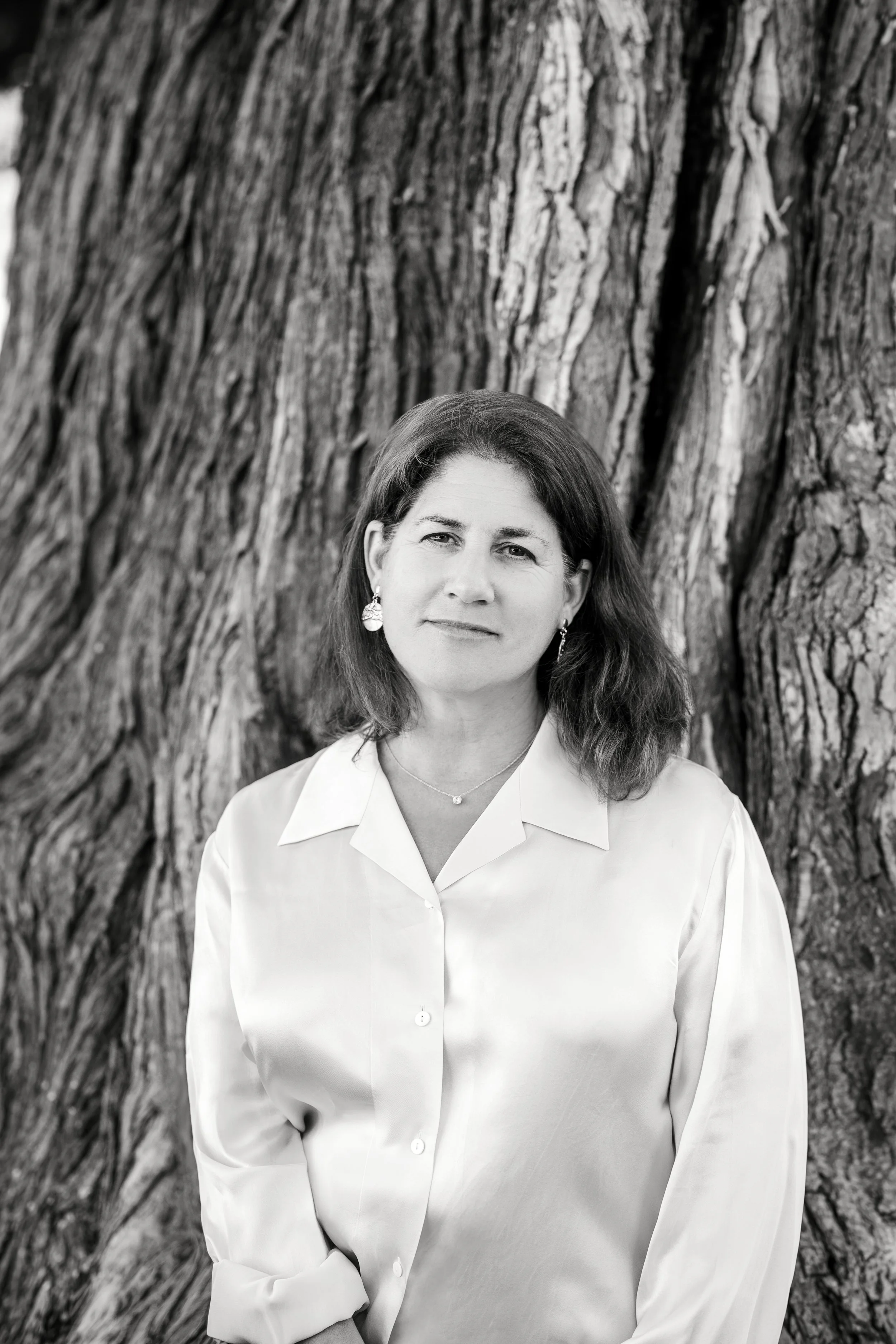 A black-and-white portrait of a woman with shoulder-length hair, wearing a collared shirt, standing in front of the textured trunk of a large tree.