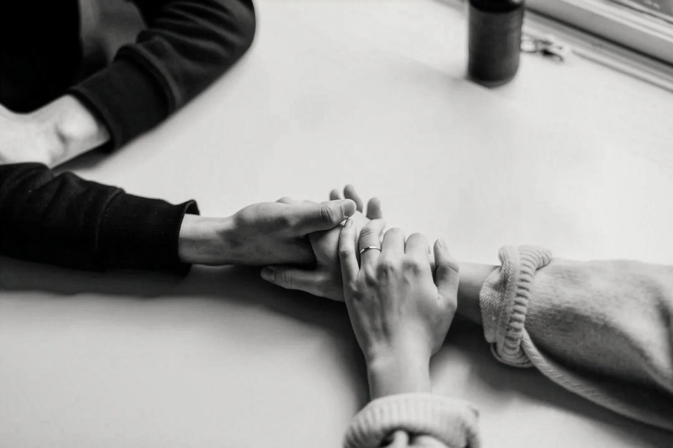 Close-up of a hand resting on a table, with another hand touching it. The scene is in black and white, showing arms in dark and light clothing.