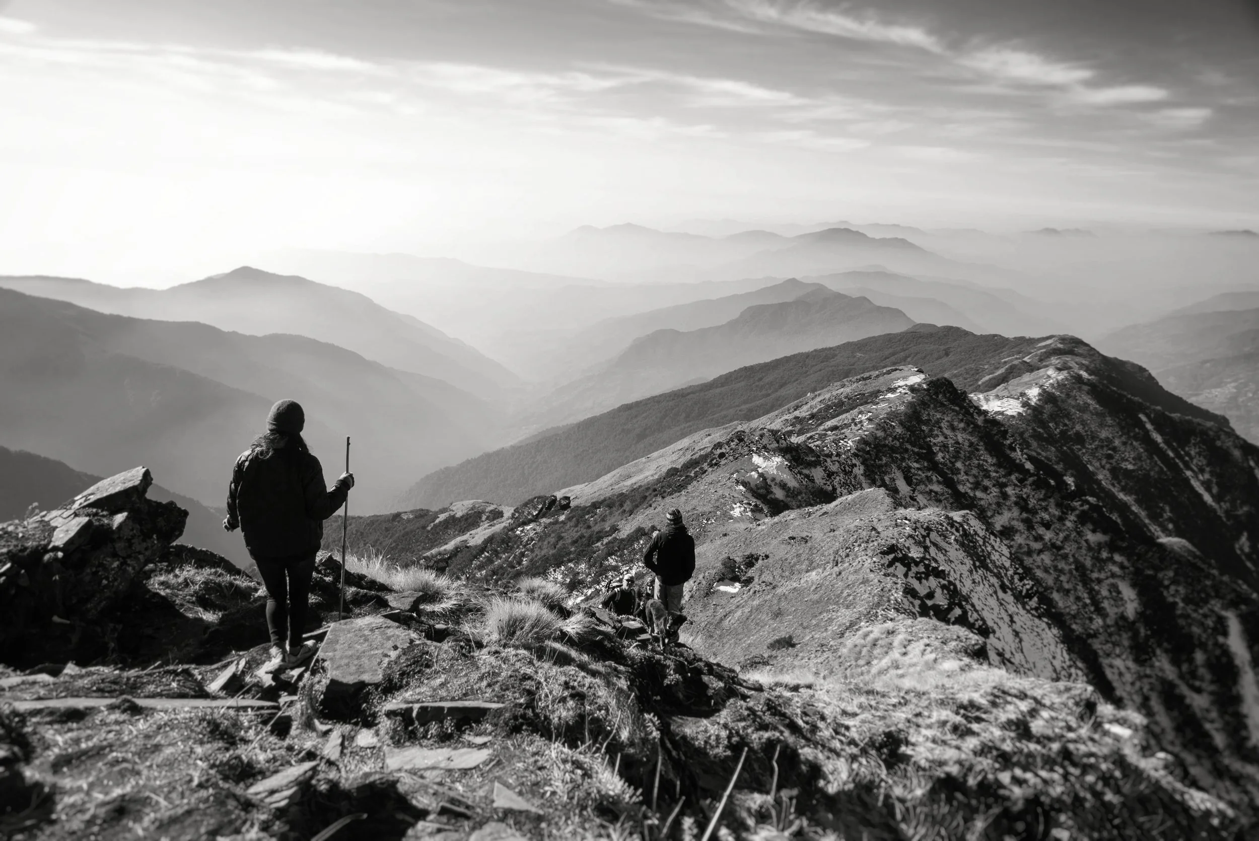 People hiking on a mountain trail with a vast mountain range in the background in black and white.