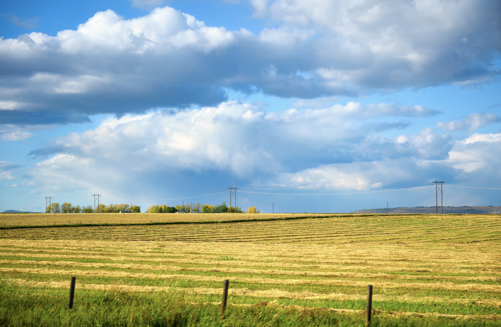 Open field with dry grass and a few posts in the foreground, and trees and hills in the background under a partly cloudy sky.