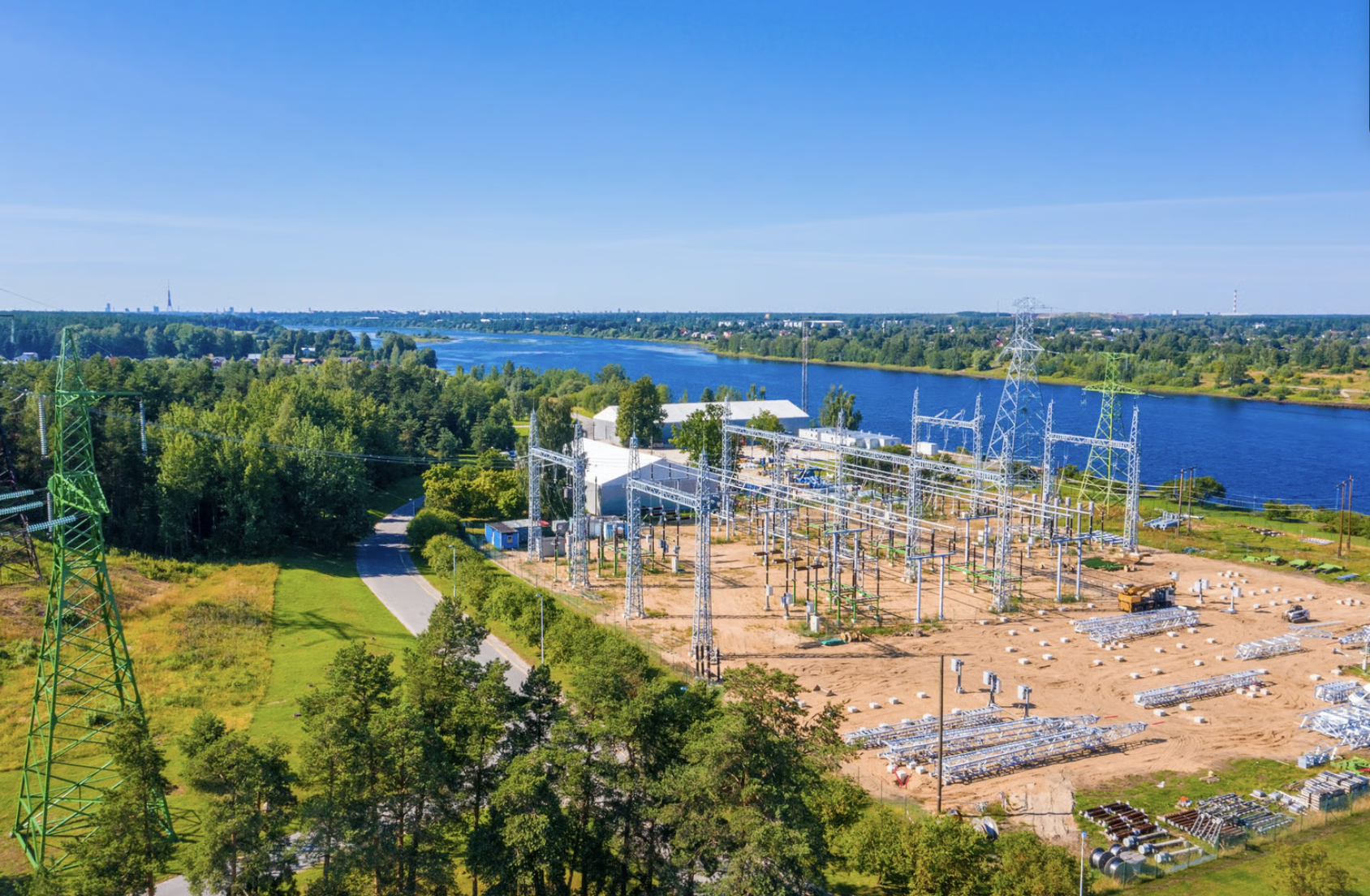 Aerial view of a power substation by a river, with trees and a path nearby under a clear blue sky.