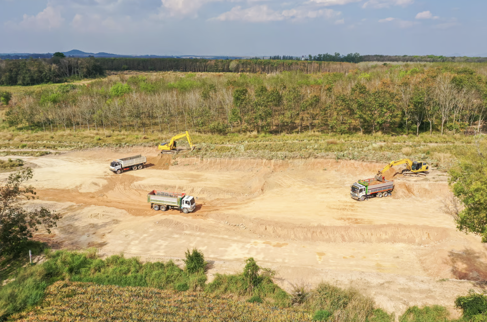 Construction site with trucks and excavators digging and moving earth, surrounded by trees and open land.