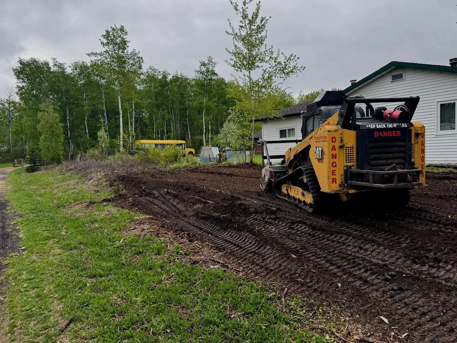 A small yellow bulldozer working on a dirt patch next to a white house with green trim, with a wooded area in the background under a gray cloudy sky.