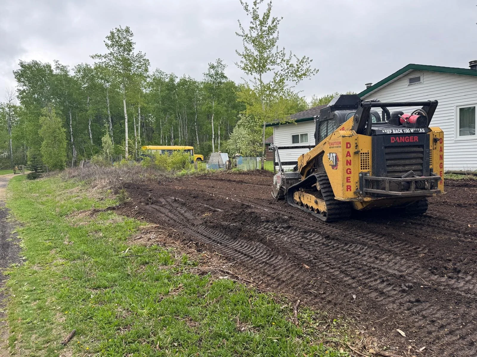 Landscaping equipment including a small bulldozer preparing a dirt area near a white house with trees in the background.
