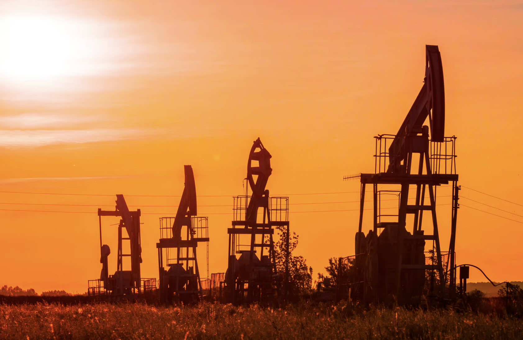 Silhouettes of oil pumpjacks against an orange sunset sky in a field.