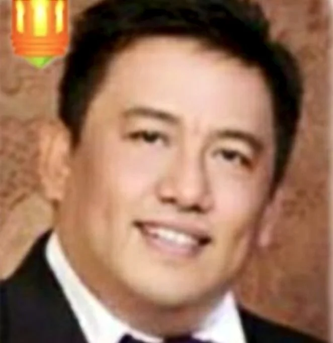 Close-up photo of a young man with short dark hair, smiling, wearing a tuxedo, with a blurred brick wall background.