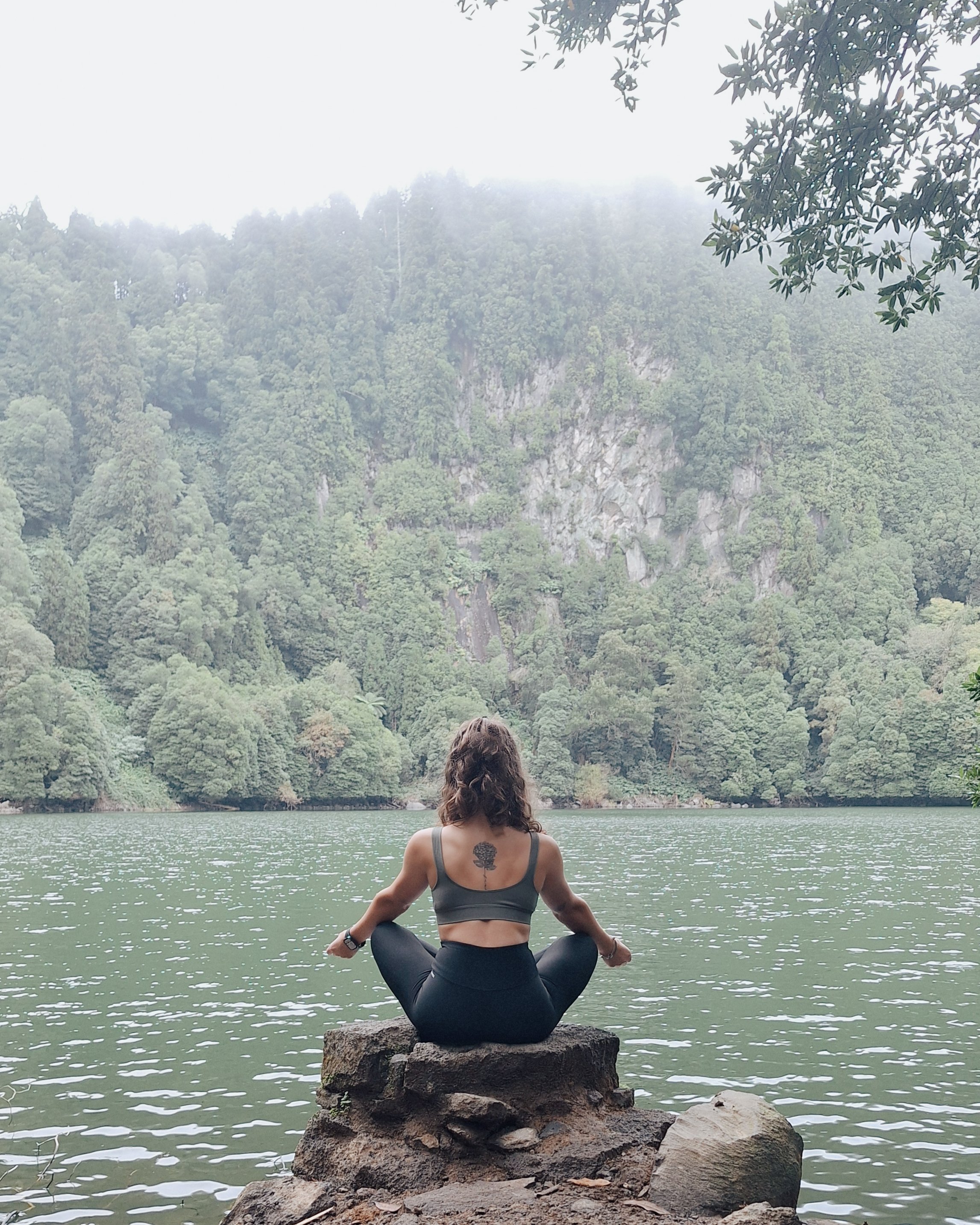 Mulher praticando yoga sentada em uma pedra às margens de um lago, com uma floresta densa ao fundo e neblina cobrindo o topo das árvores.