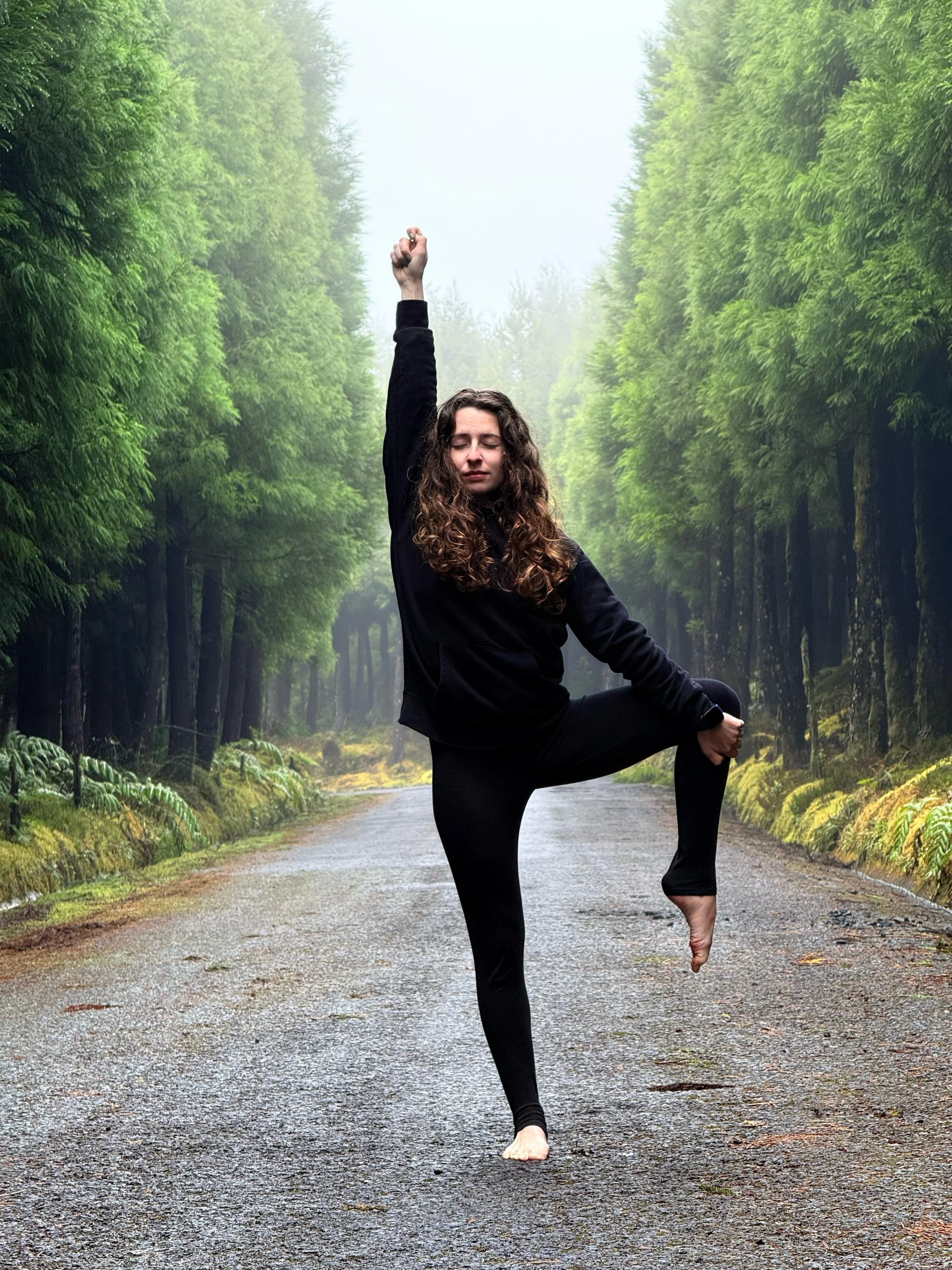 Pessoa praticando ioga na floresta, com cabelo encaracolado, usa roupas pretas, em pose de equilíbrio na estrada de terra rodeada por árvores verdes